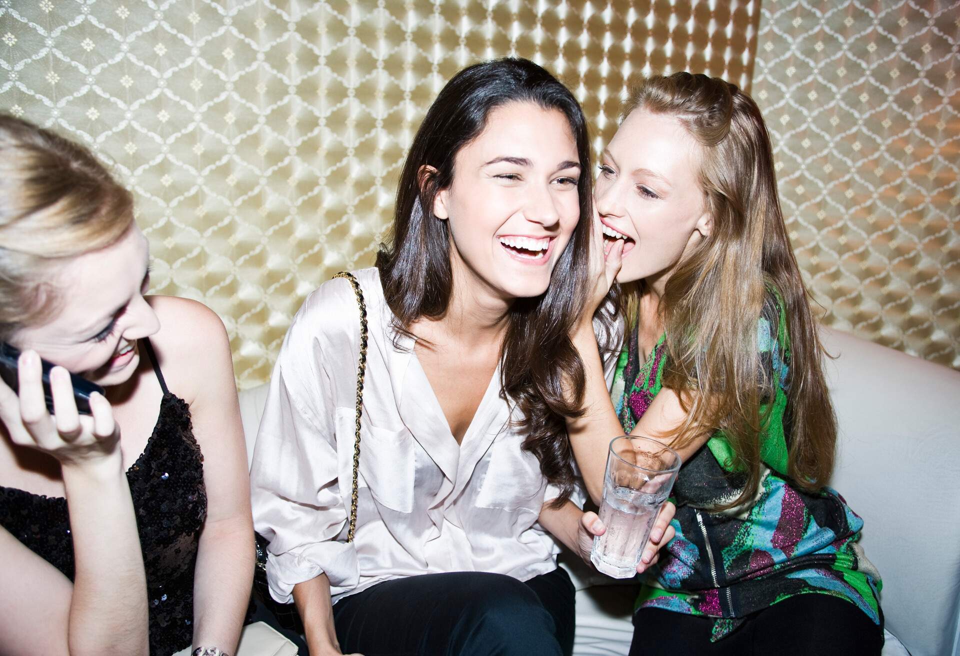 A woman whispers to a smiling friend as they sit and have drinks in a bar.