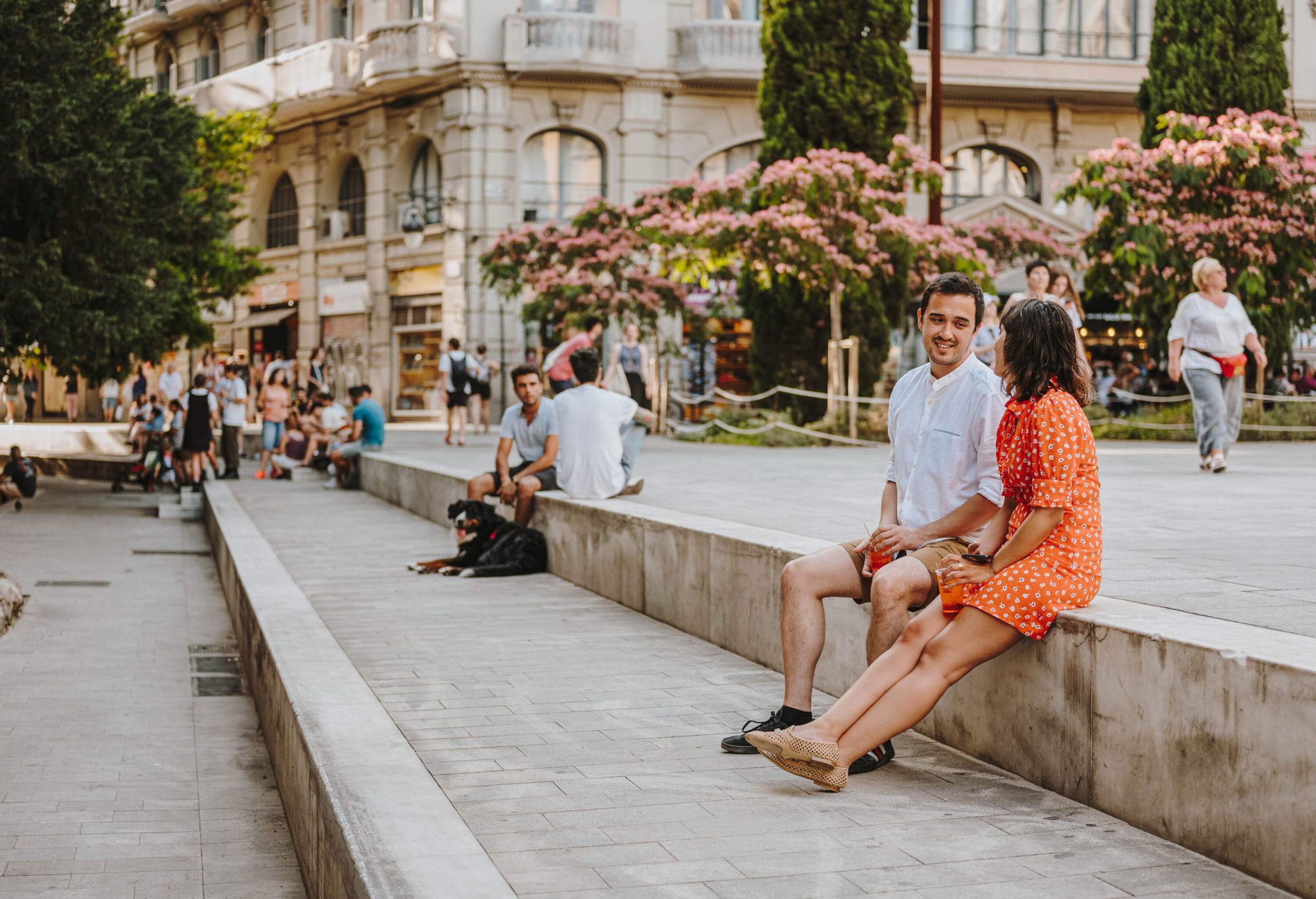 People sit leisurely on a plaza while vibrant blooming trees and charming buildings provide a picturesque backdrop.