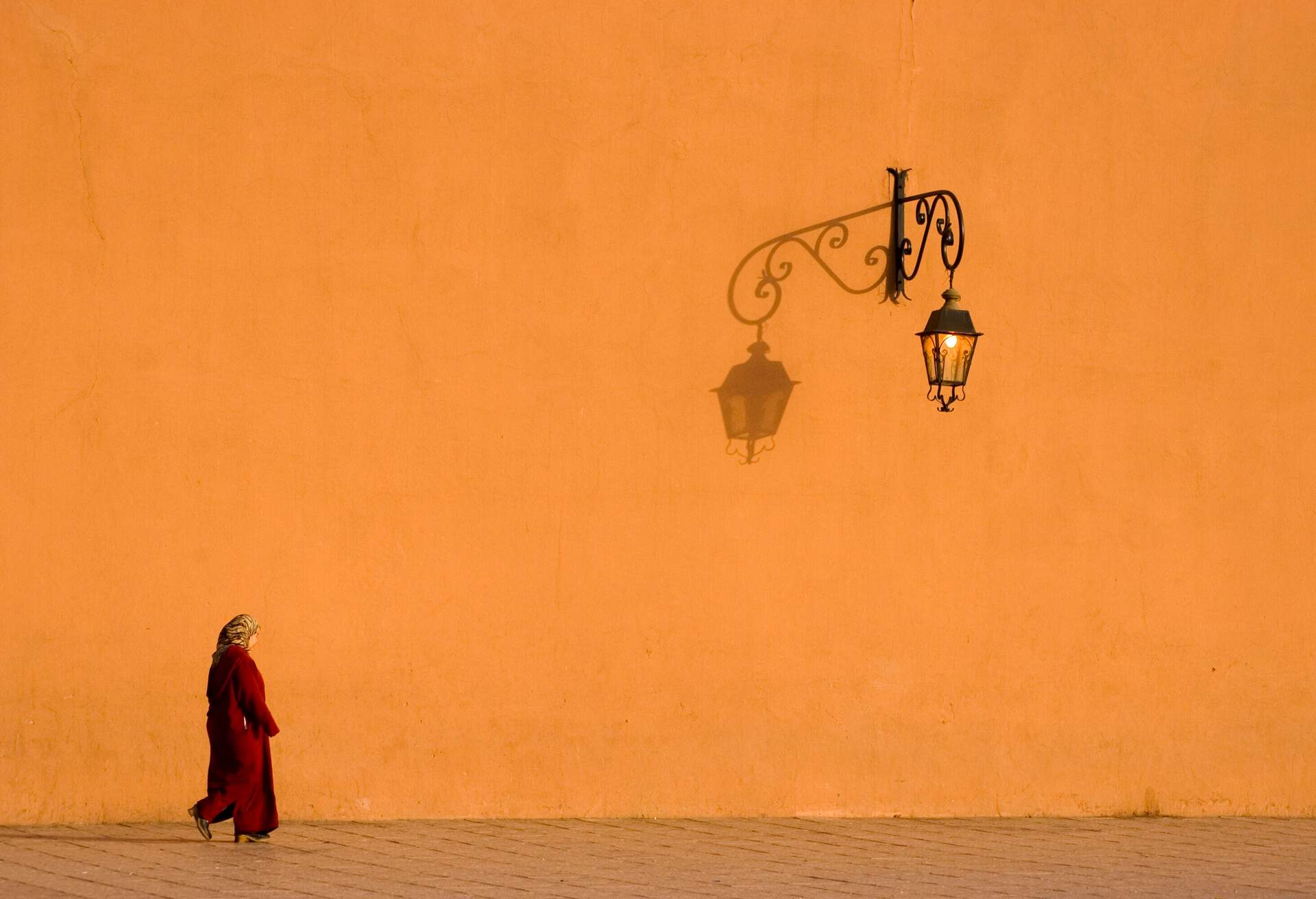A woman in a traditional red dress walks on a street against the orange wall.