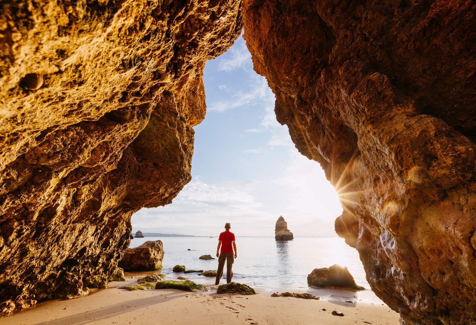 A man in a red shirt stands in the middle of the cave mouth enjoying the sunrise.