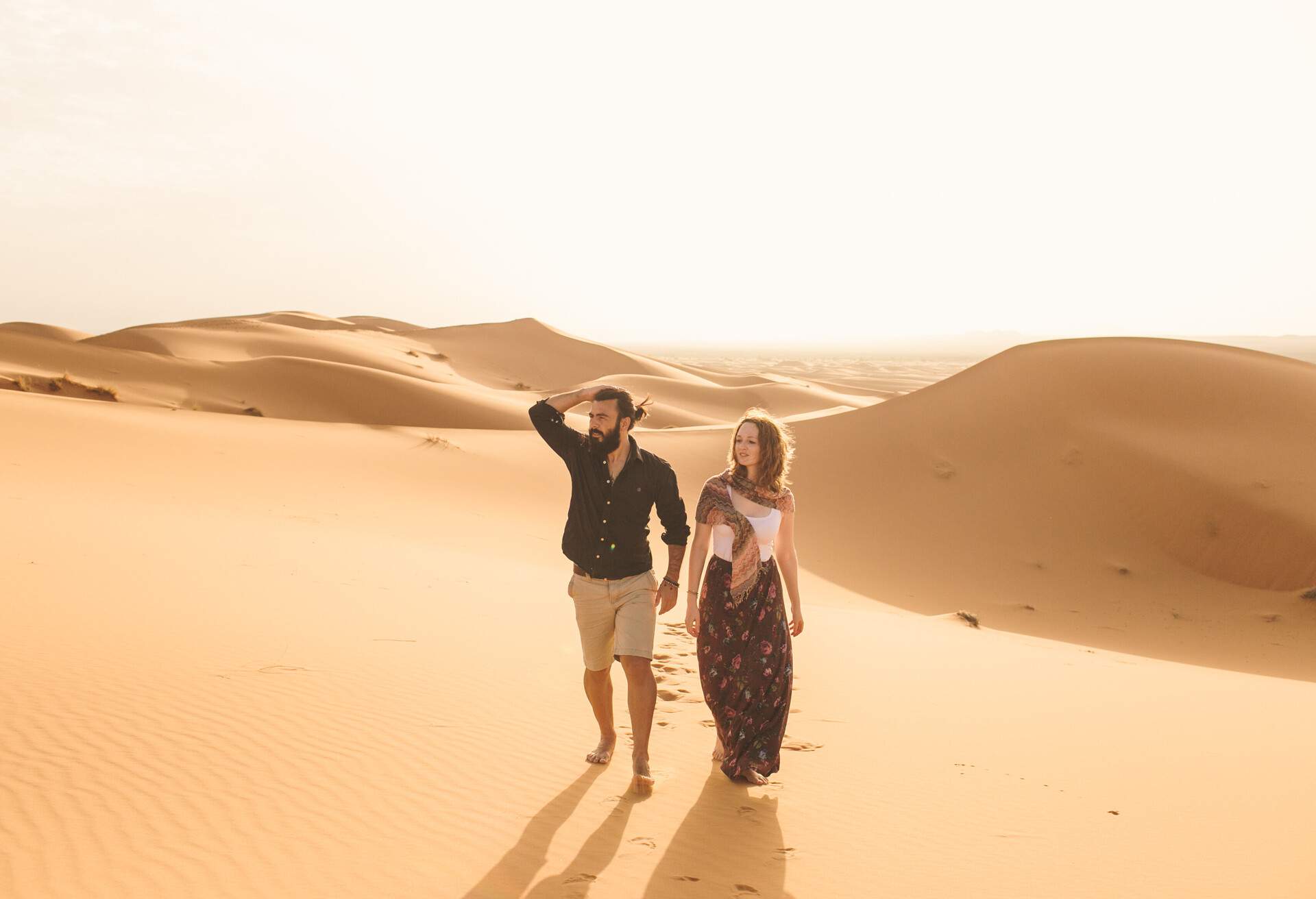 Two people strolling down a sand dune in casual dress against a blue sky.