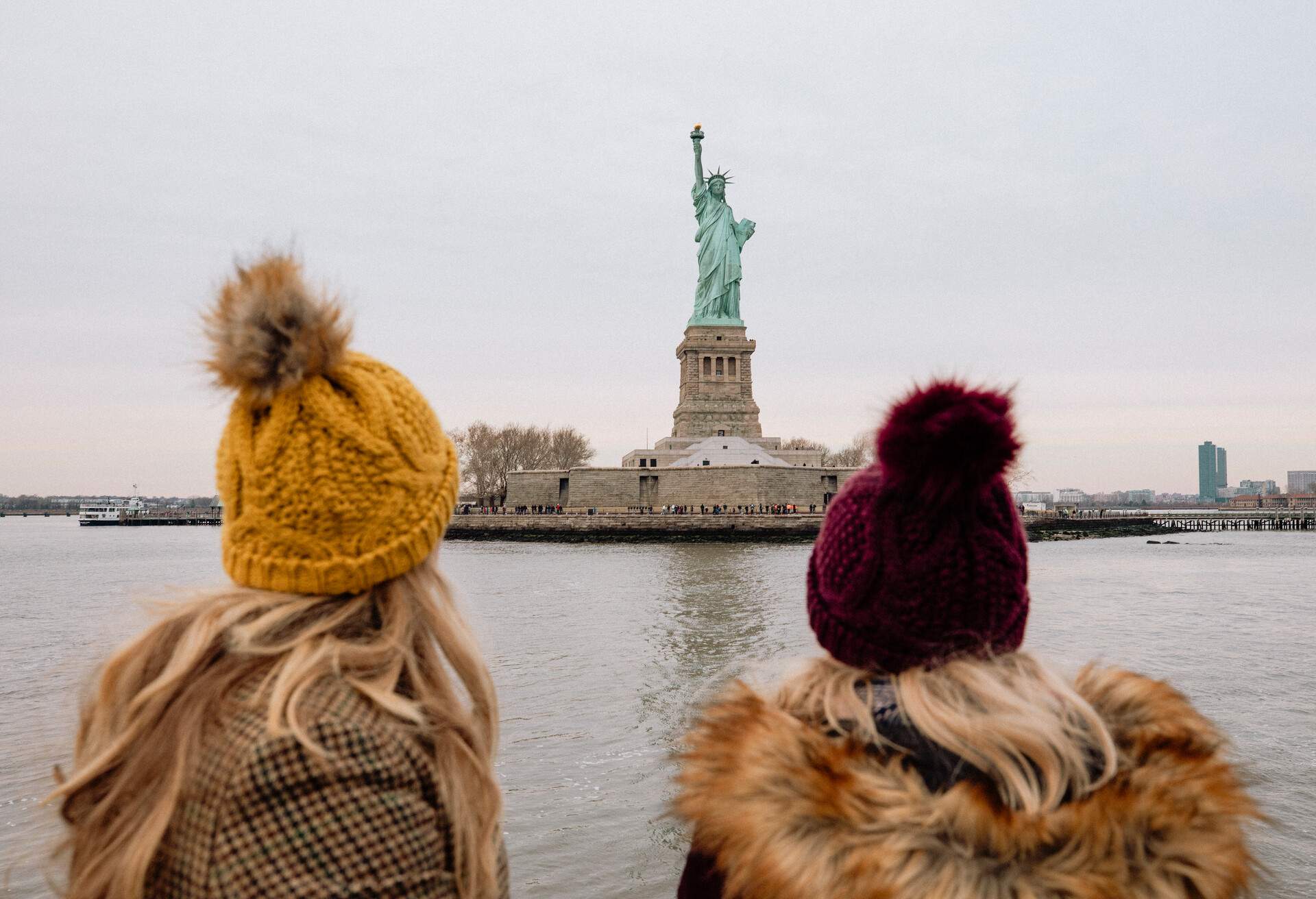 Two blonde women in black and yellow bonnet stare at the Statue of Liberty on an island.