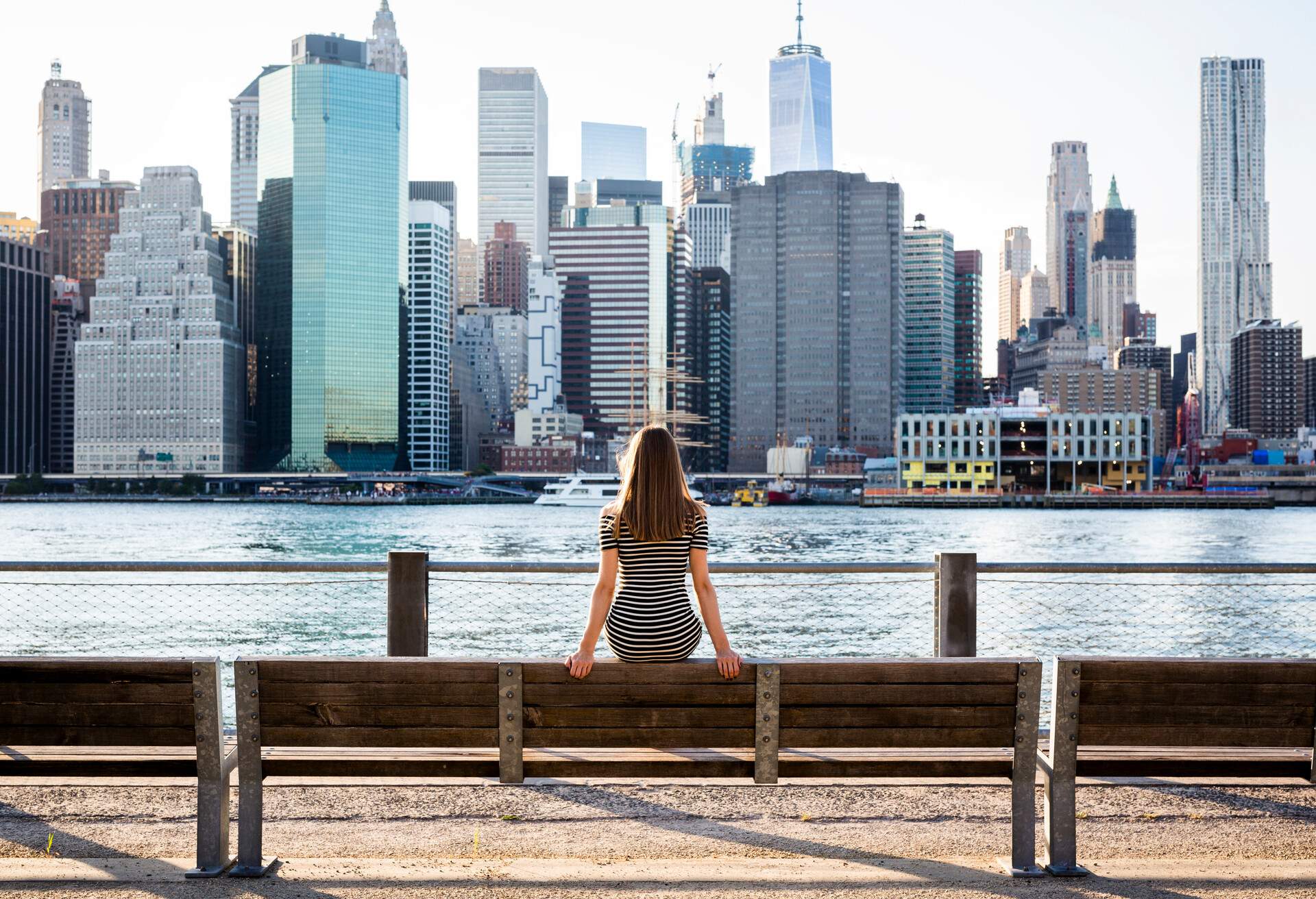 A woman sitting on the backrest of a bench seat facing the massive buildings across a river.