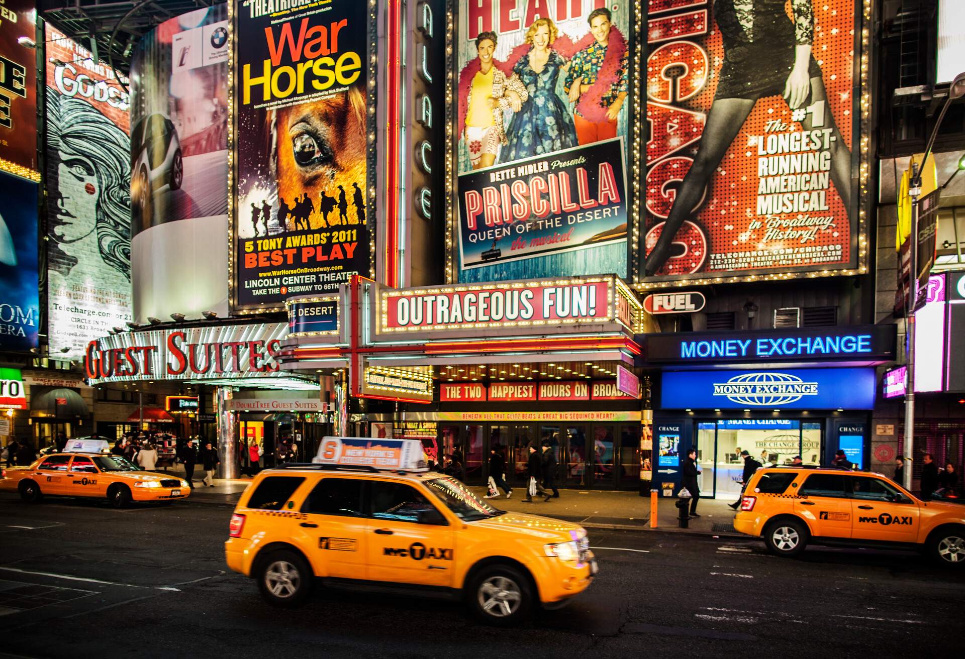 Iconic Broadway theatres in the heart of Times Square, New York City, come alive with dazzling poster ads, as yellow taxis in the foreground add a vibrant touch to the bustling scene.