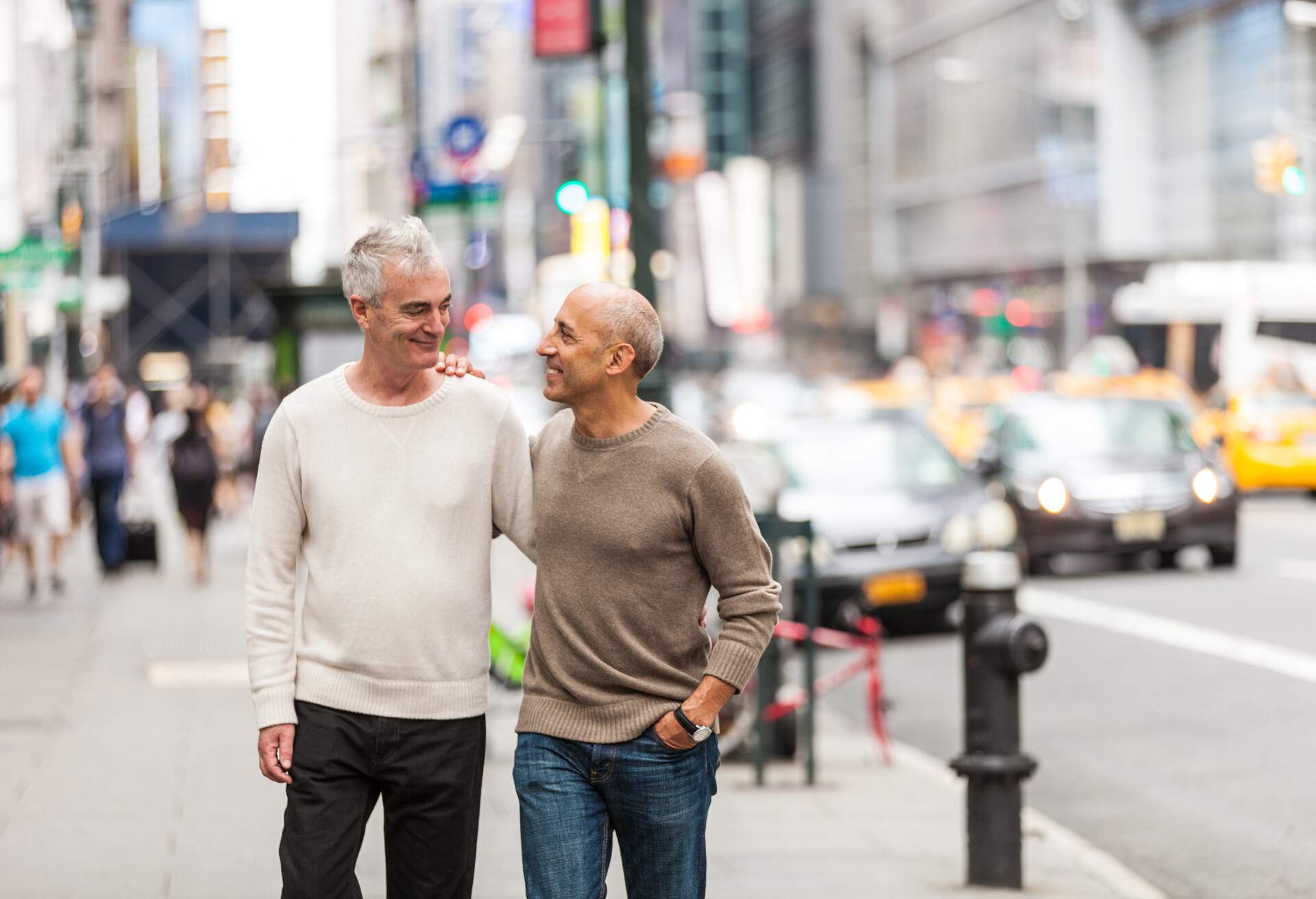A male couple standing on the side walk of a bustling city and looking fondly at each other.