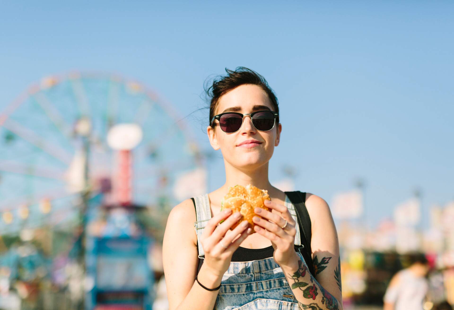 A tattooed young woman standing in an amusement park holding a sandwich with a big Ferris wheel behind her.