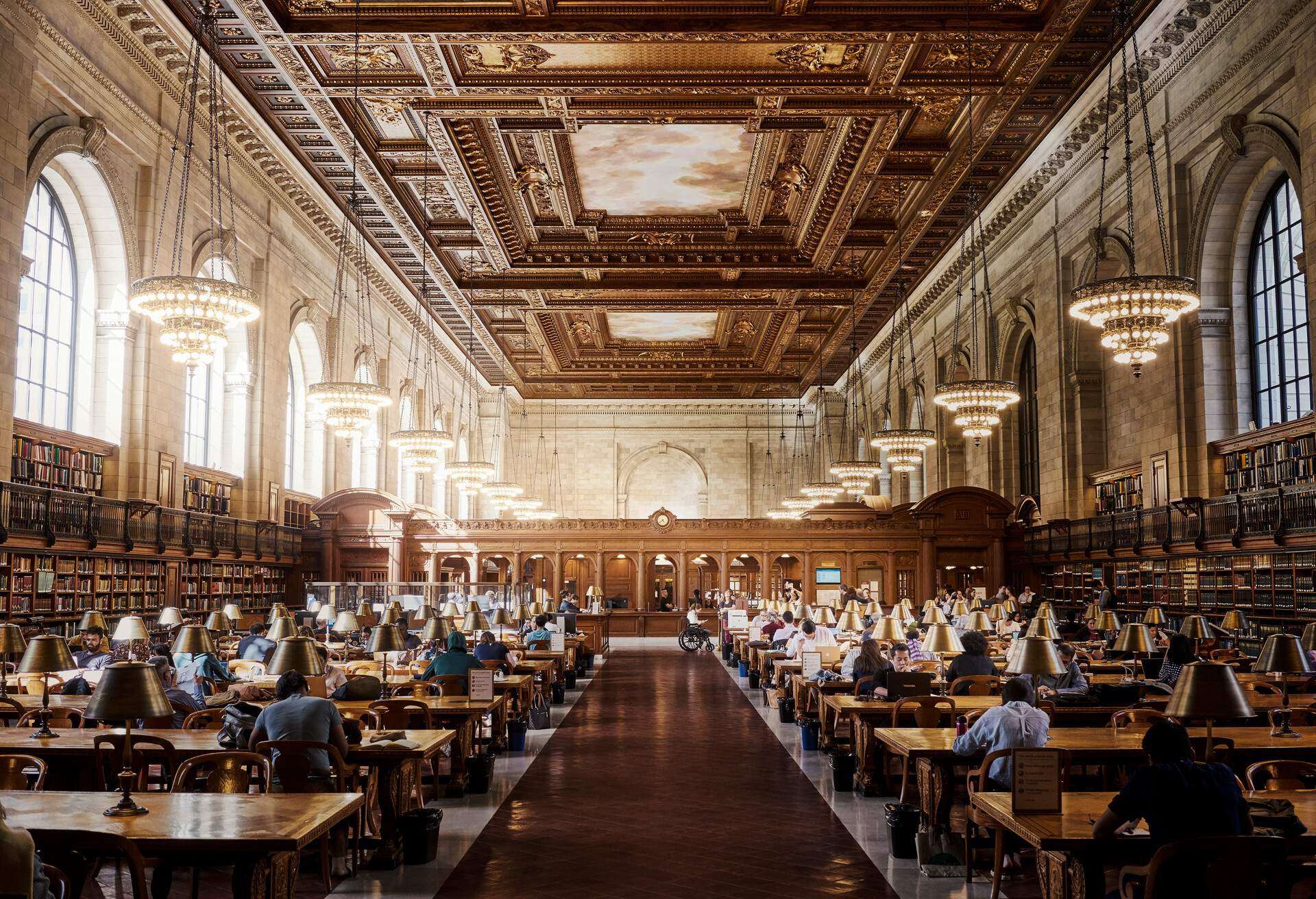 Interior of a library with intricately carved ceilings, hanging chandeliers, bookshelves and rows of tables with people studying.