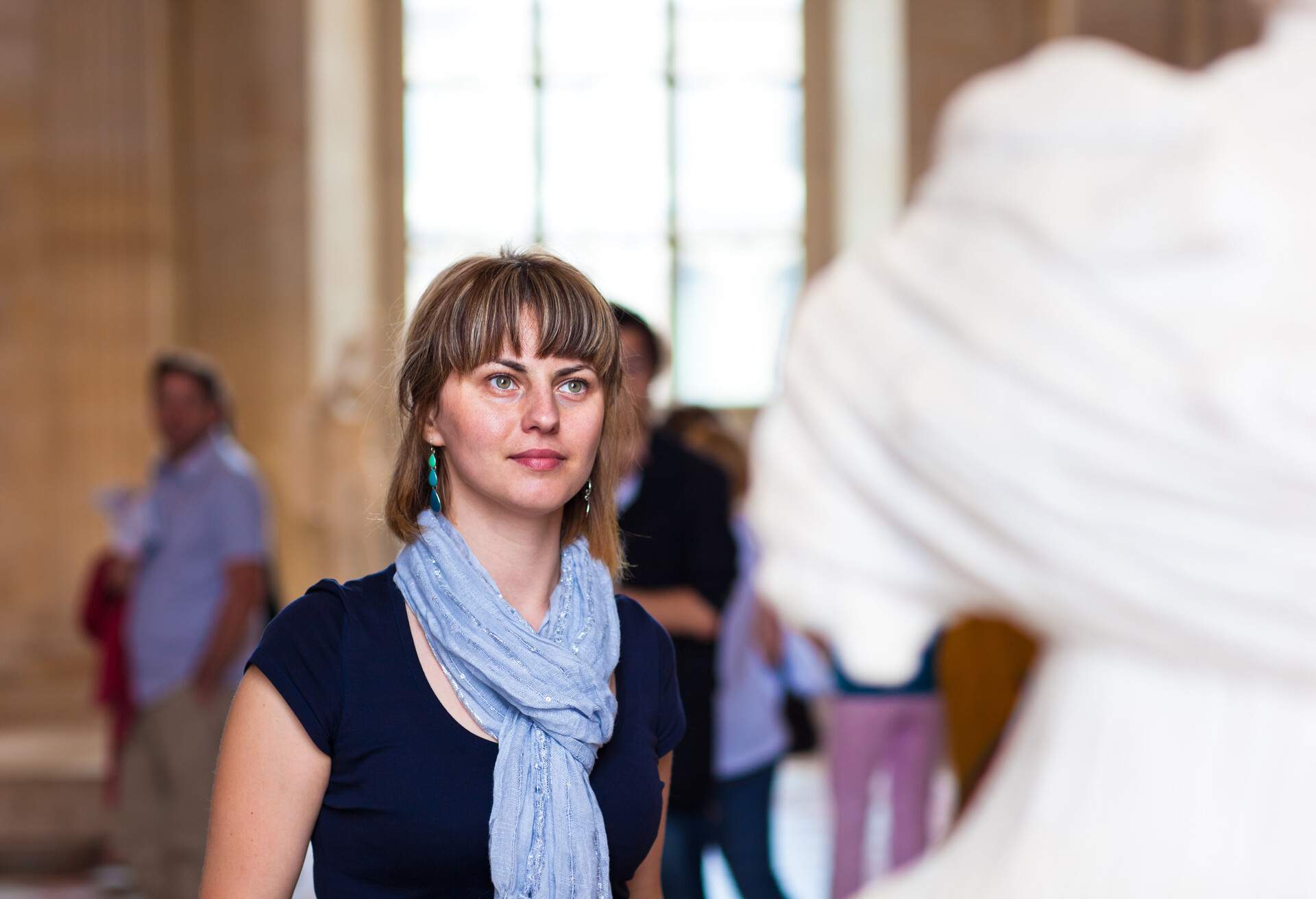 A woman wearing a light blue scarf and dark blue blouse admires the exquisite craftsmanship of a fine art statue, fully immersed in the museum experience.