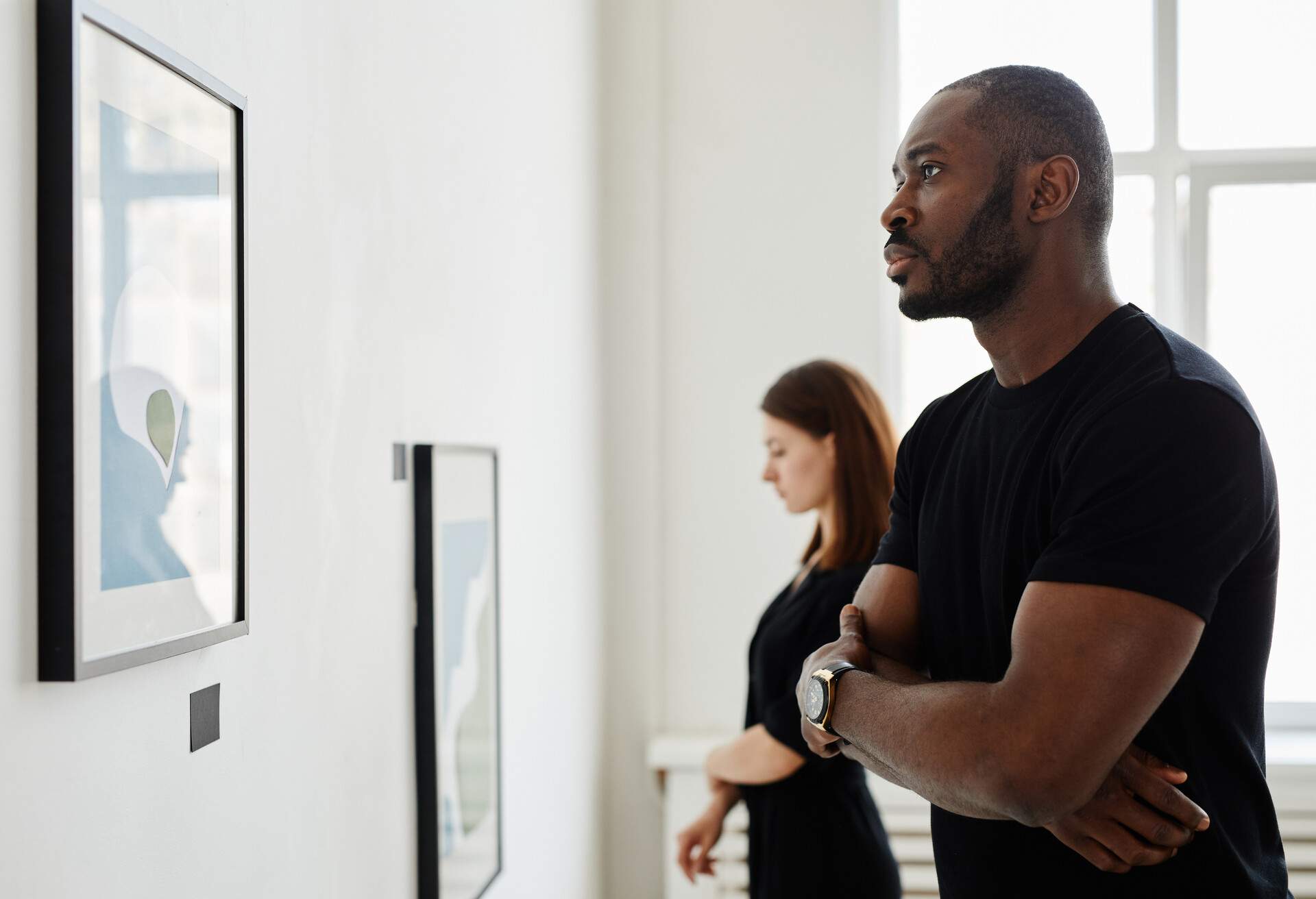 A man in a black shirt thoughtfully examines paintings in a modern art gallery, while another person in the background is engrossed in a different artwork.