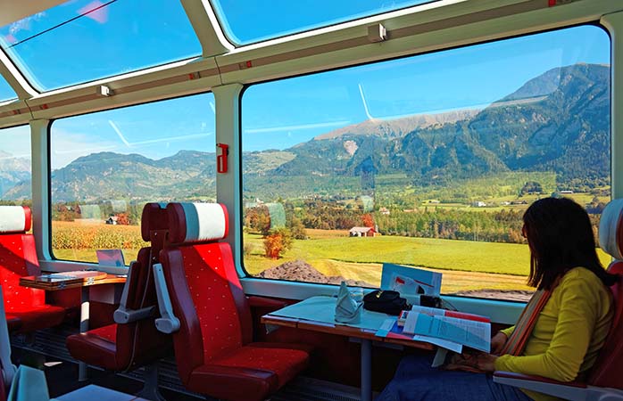 A passenger enjoying the idyllic scenery of Swiss countryside through the wide panoramic windows on the Glacier Express with sunlight cast thru the glass skylights on a sunny summer day in Switzerland; Shutterstock ID 751719448