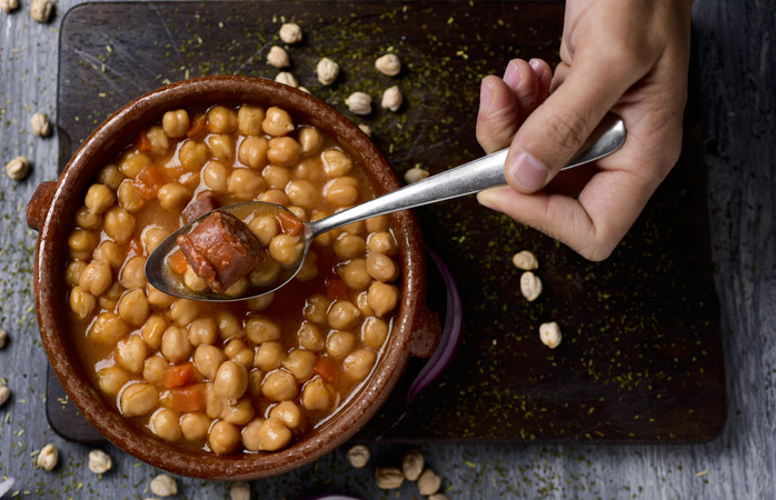 high-angle shot of the hand of a man with a spoon and an earthenware bowl with spanish cocido madrileno, a stew typical of madrid with chickpeas, pork, bacon, chorizo and ham, on a rustic wooden table