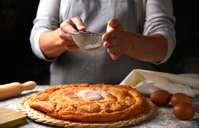 closeup of a young caucasian man sprinkling powdered sugar on an ensaimada, a pastry typical of Mallorca, Spain, placed on a rustic wooden table; Shutterstock ID 1036889518