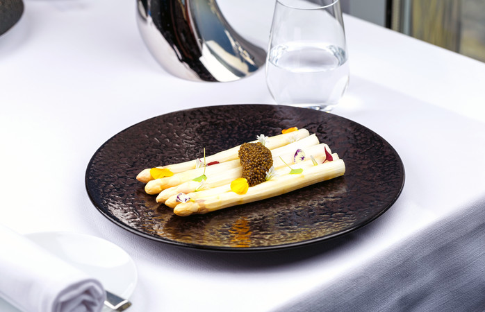 White asparagus served with caviar decorated with flowers on black plate on white tablecloth.