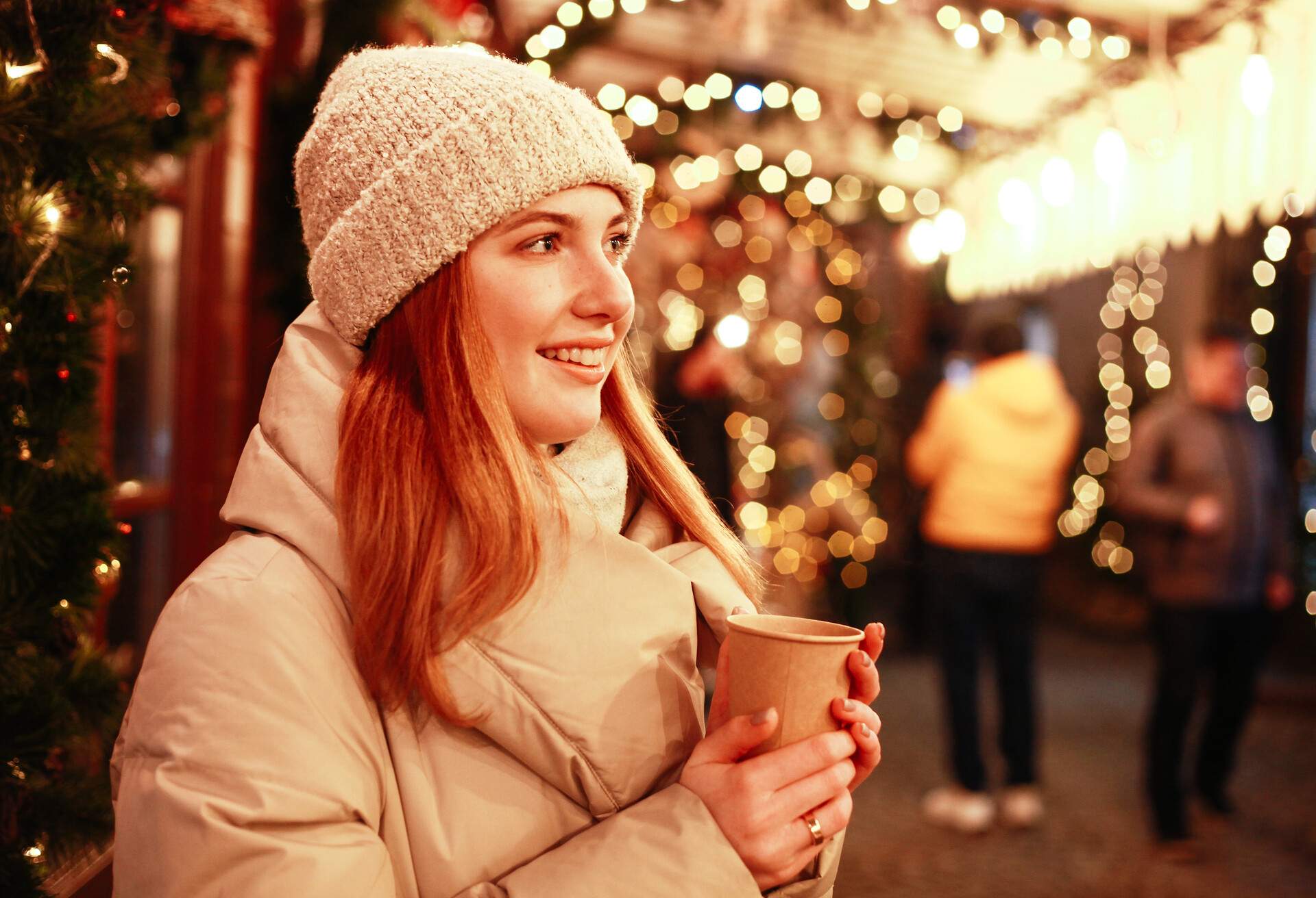 A woman in winter clothes is looking away, smiling and holding a drink at the illuminated Christmas market.