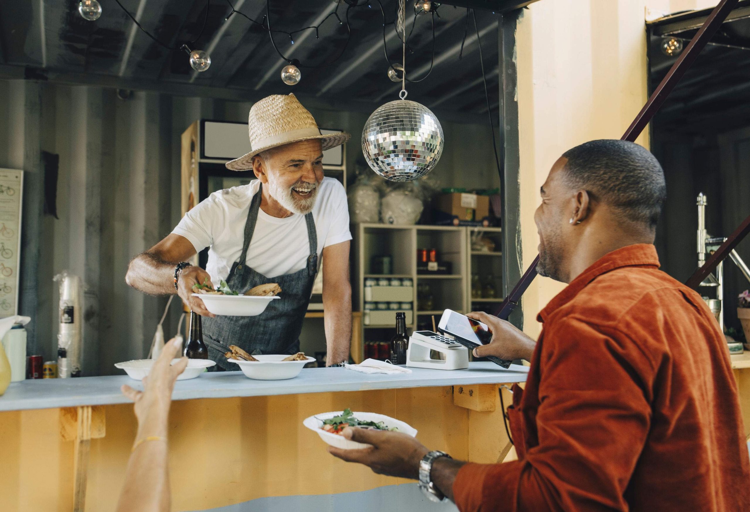 An elderly man in a straw hat serves food to a man in a brown shirt in the food truck counter.