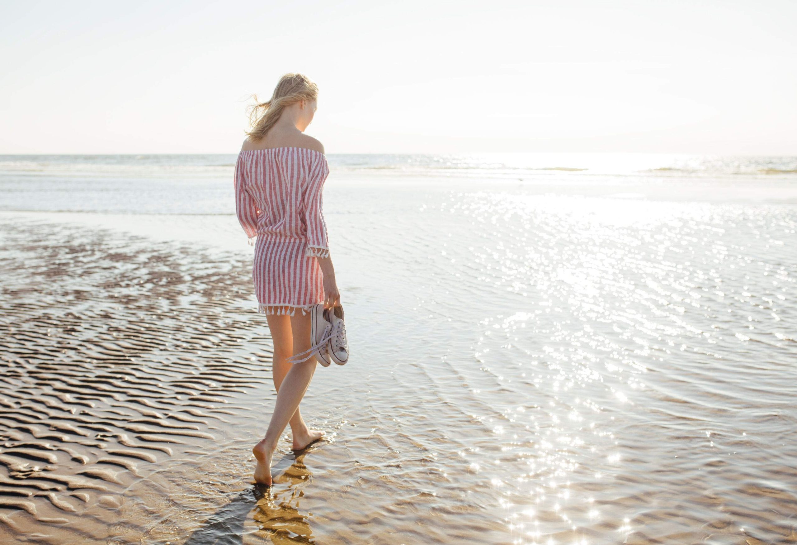 A woman walking on the beach with her sneakers in hand as the sea shimmers in the sunlight.