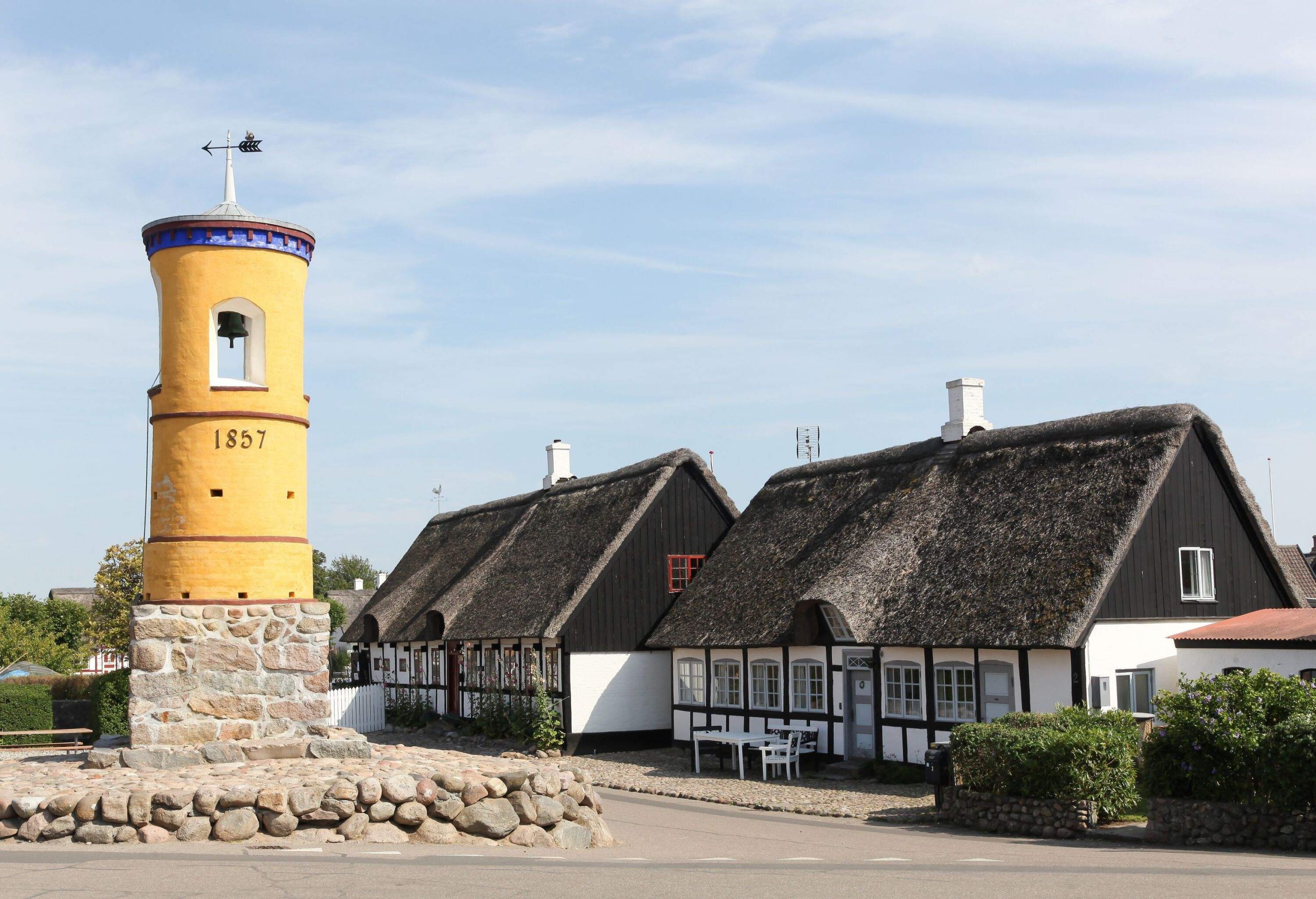 A yellow bell tower built on a drystone pedestal in front of traditional Danish farm houses.