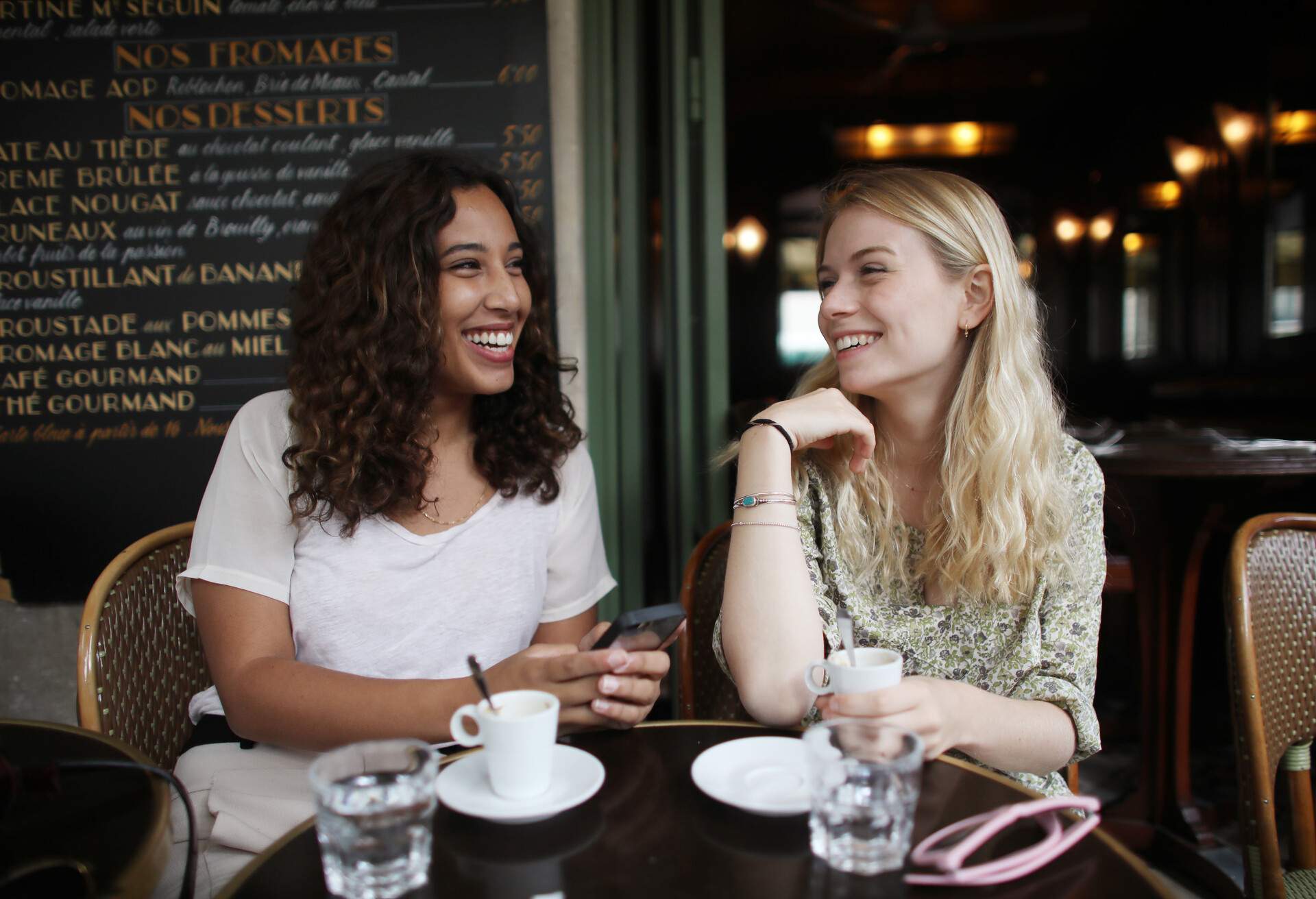 Two female friends smiling at each other as they sit in a café.