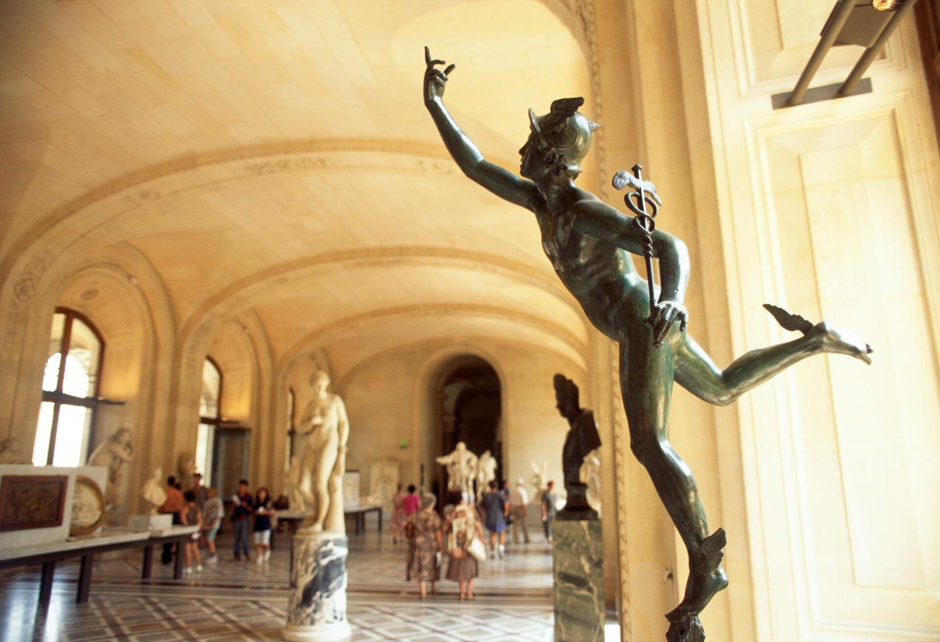 Statue of the Greek god Mercury with a winged helmet, standing on one leg and holding a rod encircled by two snakes and tipped with wings, with the other hand pointing upward against a blurred background of people and other sculptures.