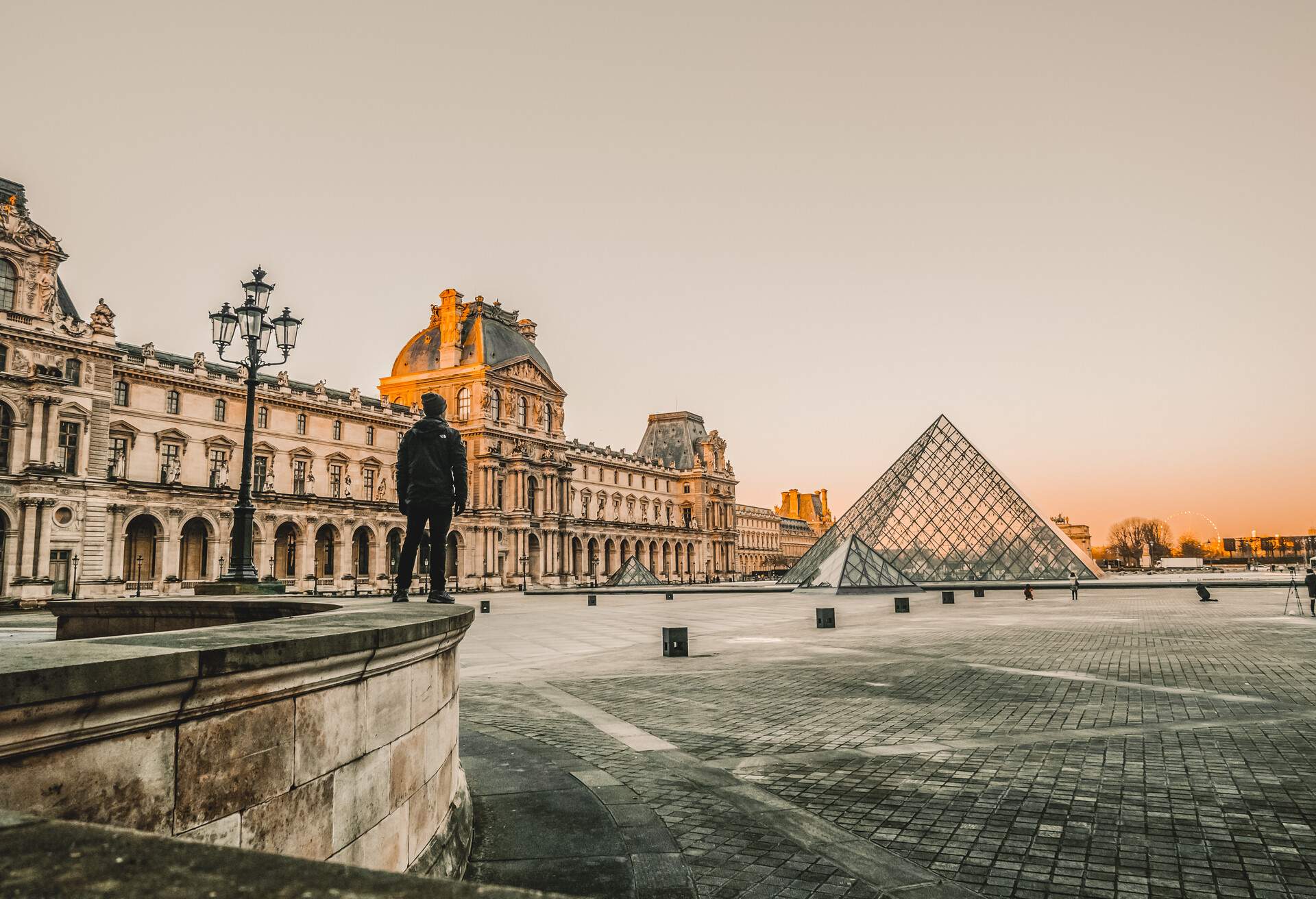A man standing on a low wall overlooking the glass pyramids of the Louvre Museum.
