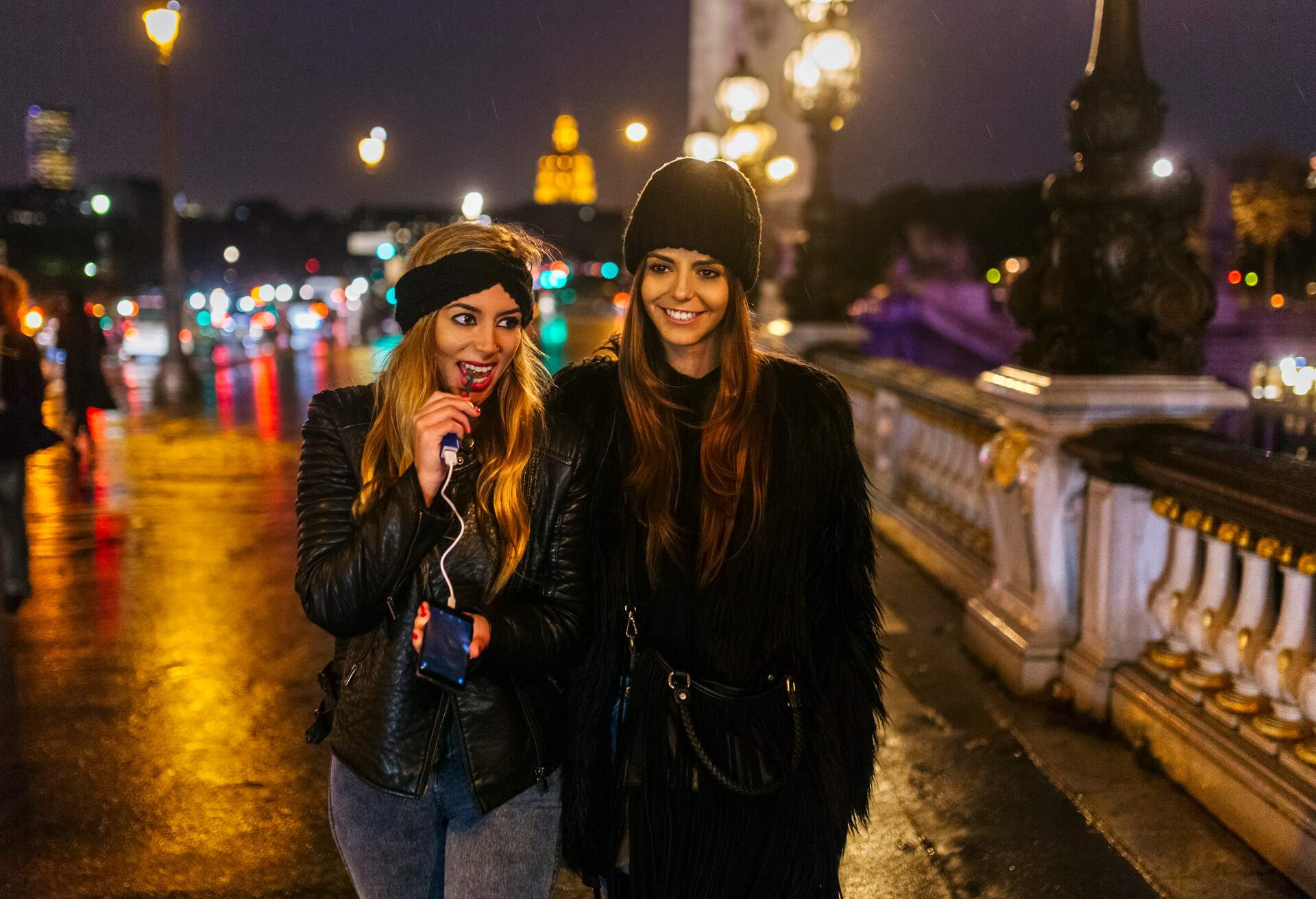 Two women dressed in black winter attire walk on a bridge with a blurred, damp background.
