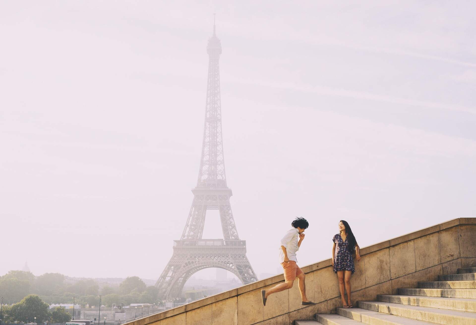 A woman leans on the staircase rails as a man skips on the steps, with a hazy view of the Eiffel Tower in the background.