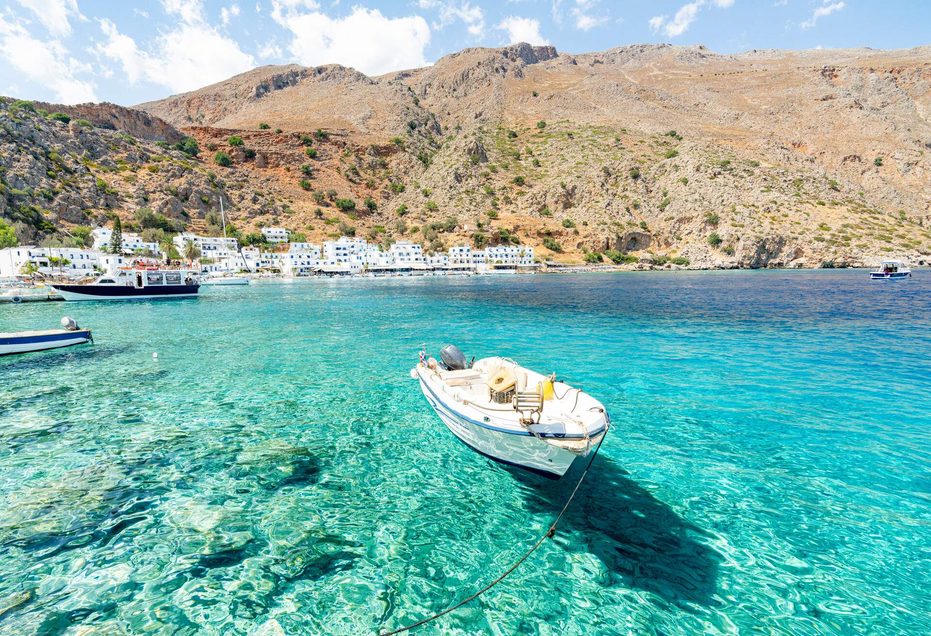 A small white boat moored on crystalline turquoise waters on a white seaside village at the base of a harsh rocky mountain.