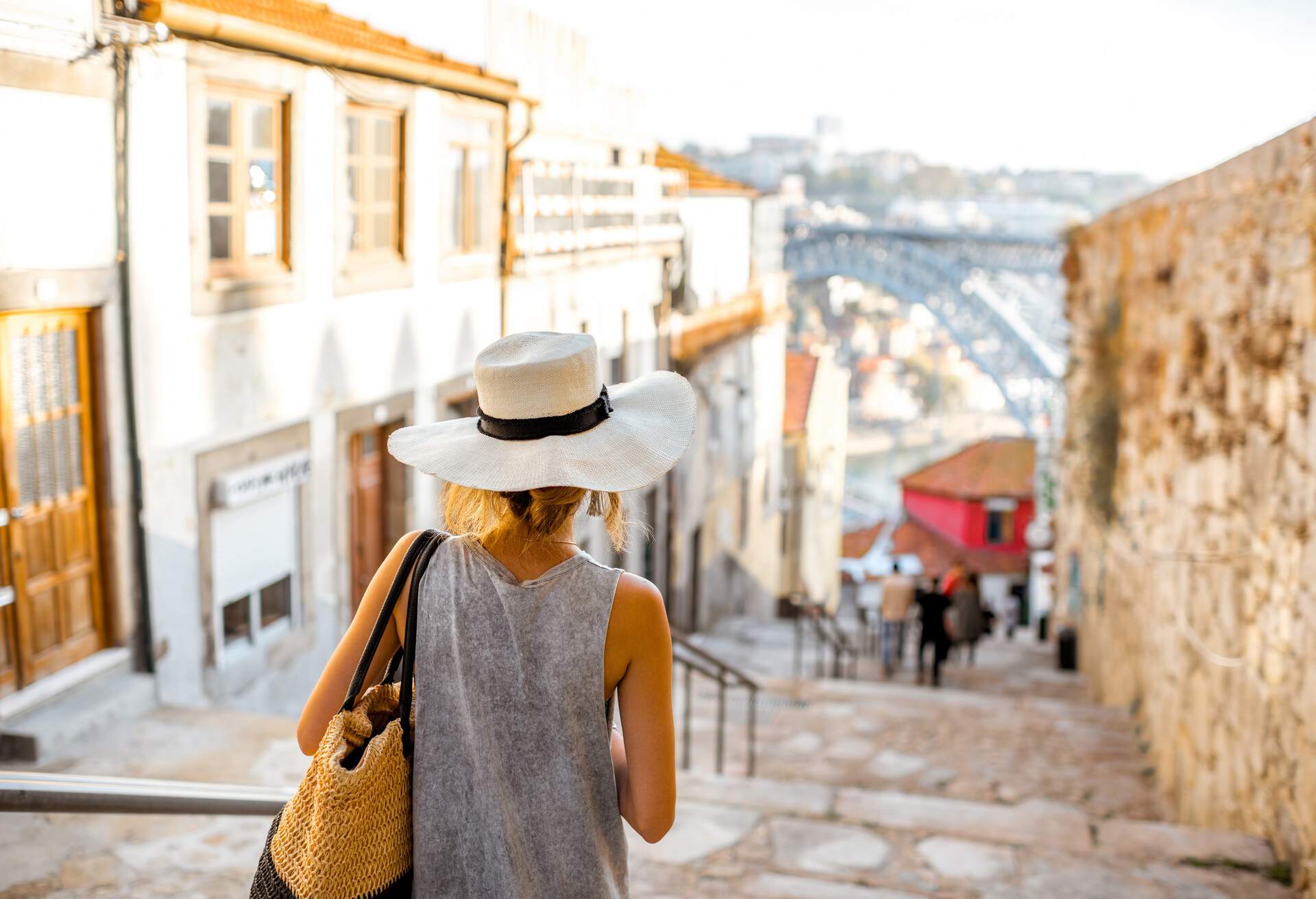 A young woman tourist, stylishly dressed and wearing a sunhat, confidently walks down the stairs of a charming, sunlit street with an out-of-focus background.