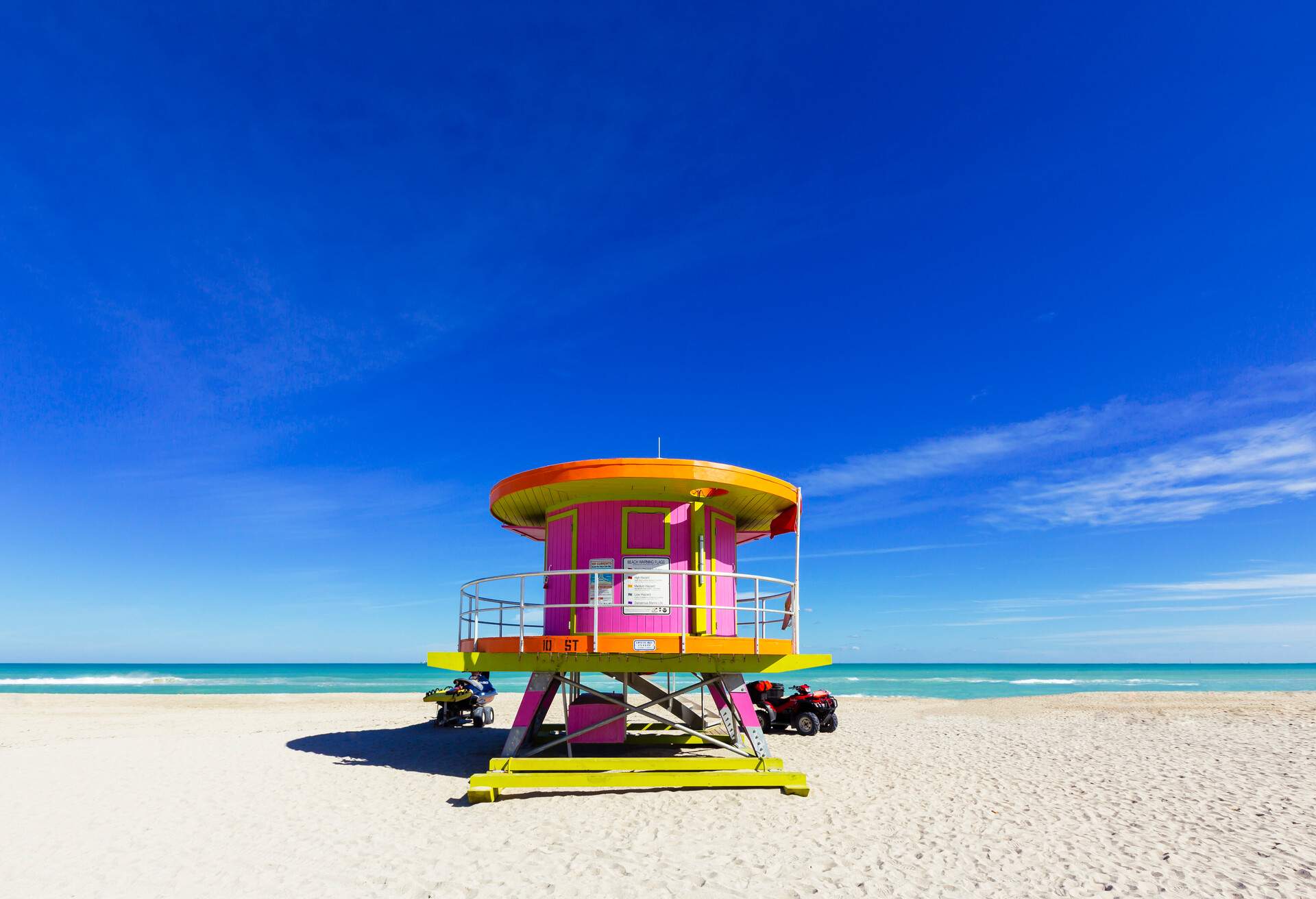 A lifeguard tower painted in vibrant colours nestled in an empty sandy beach against the clear blue sky.
