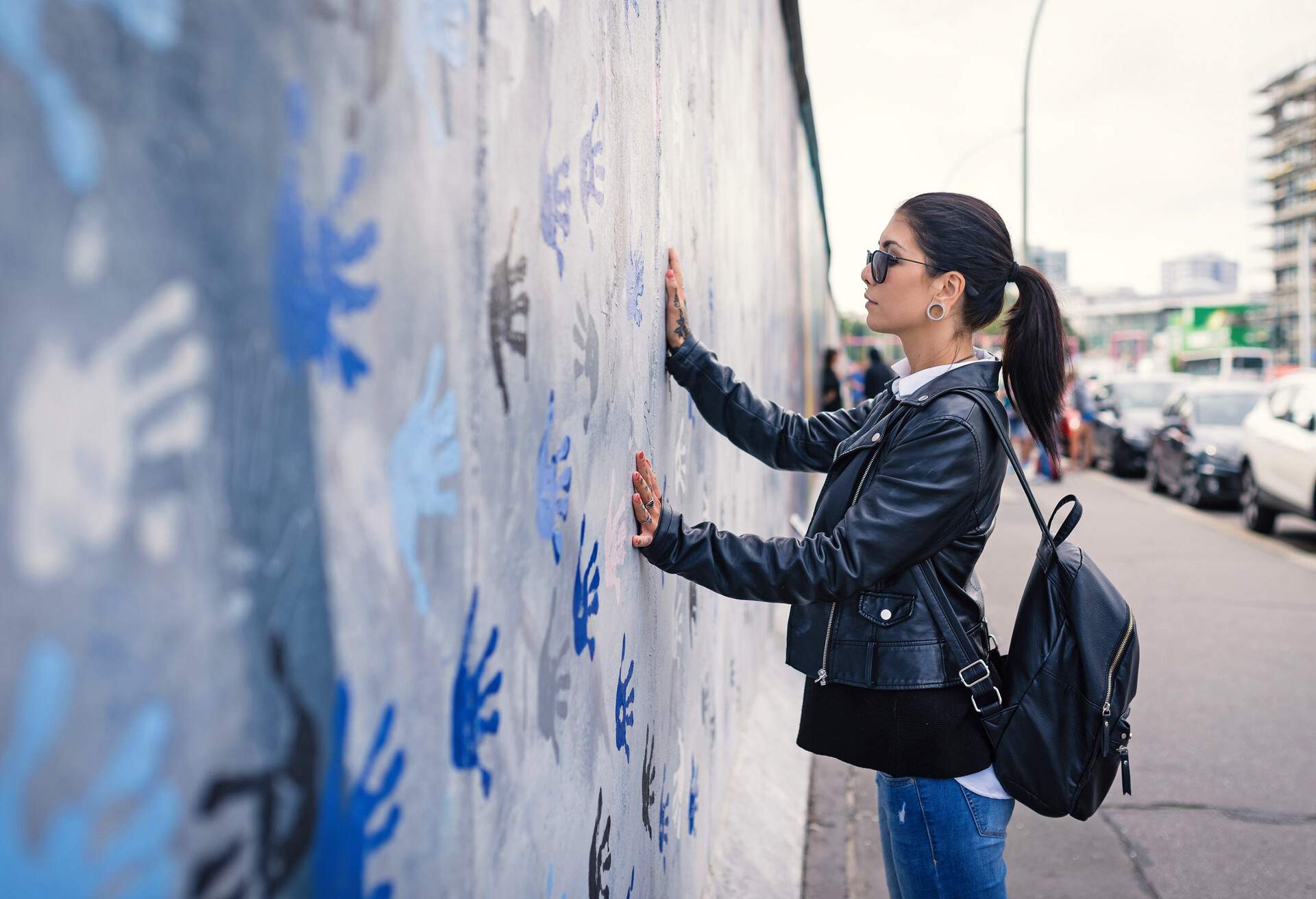 A woman in a black leather jacket stands in front of a wall decorated with colourful handprints and touches the wall with her hand.