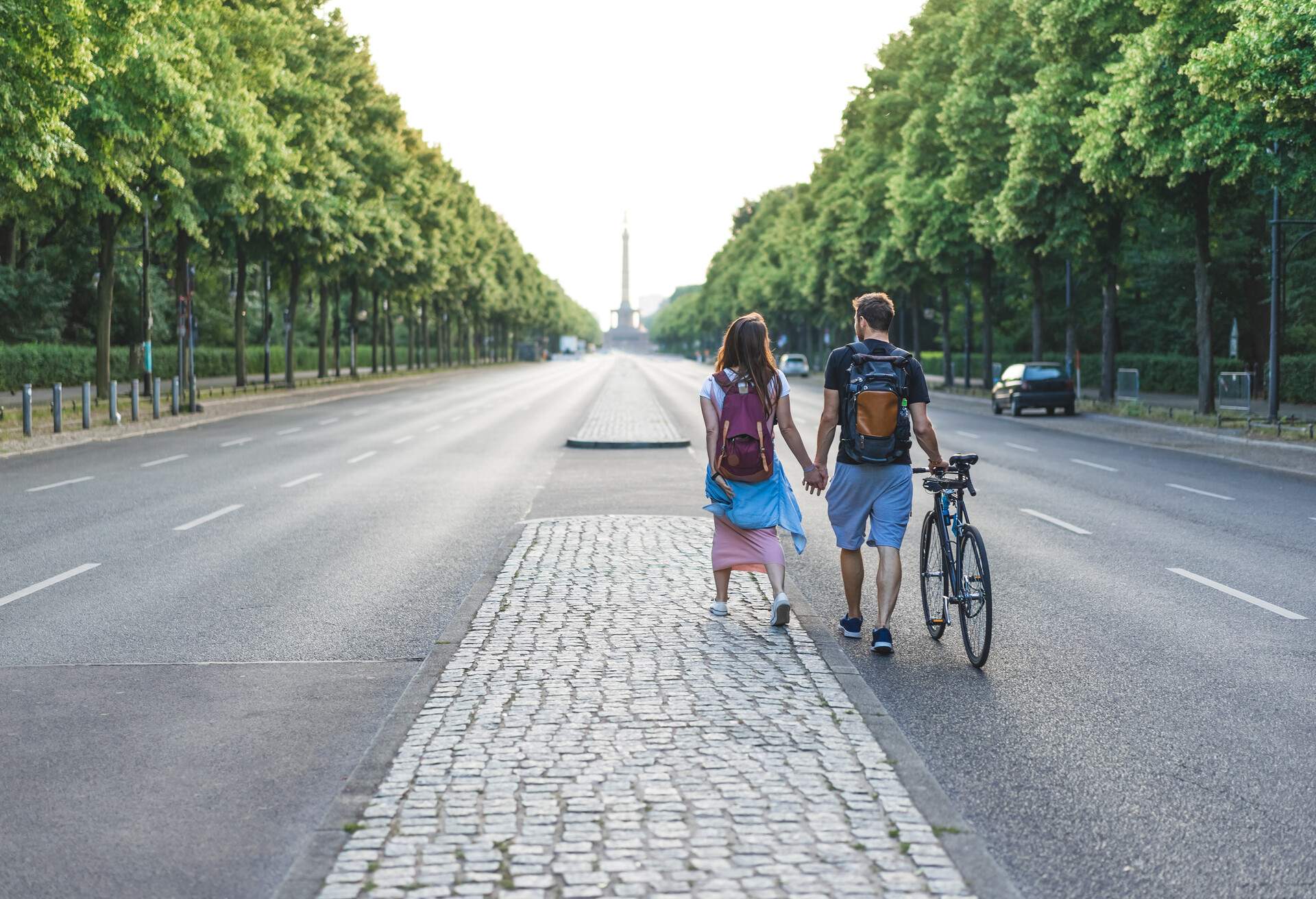 A couple with a bicycle walks hand in hand in the middle of a street lined with tall green trees.