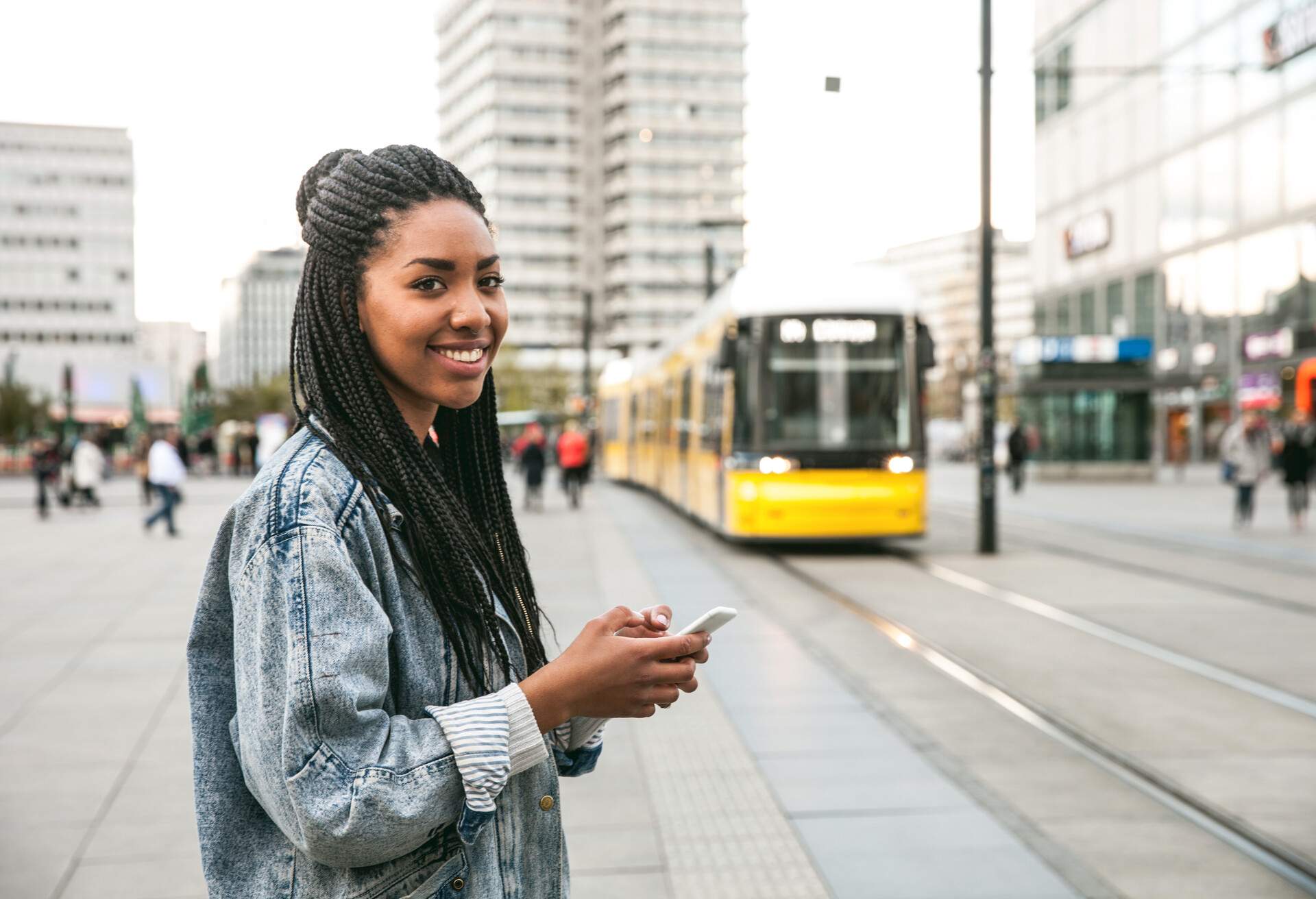 A woman with dreadlocks smiles while using her smartphone as she stands near an approaching yellow train.