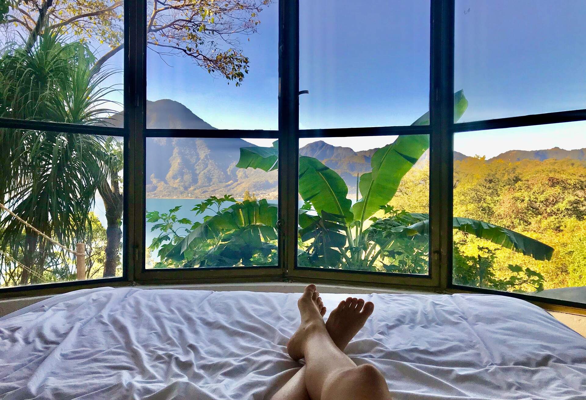 A man lying on the bed with his feet crossed at the ankles, gazing out the window at the trees and mountains.