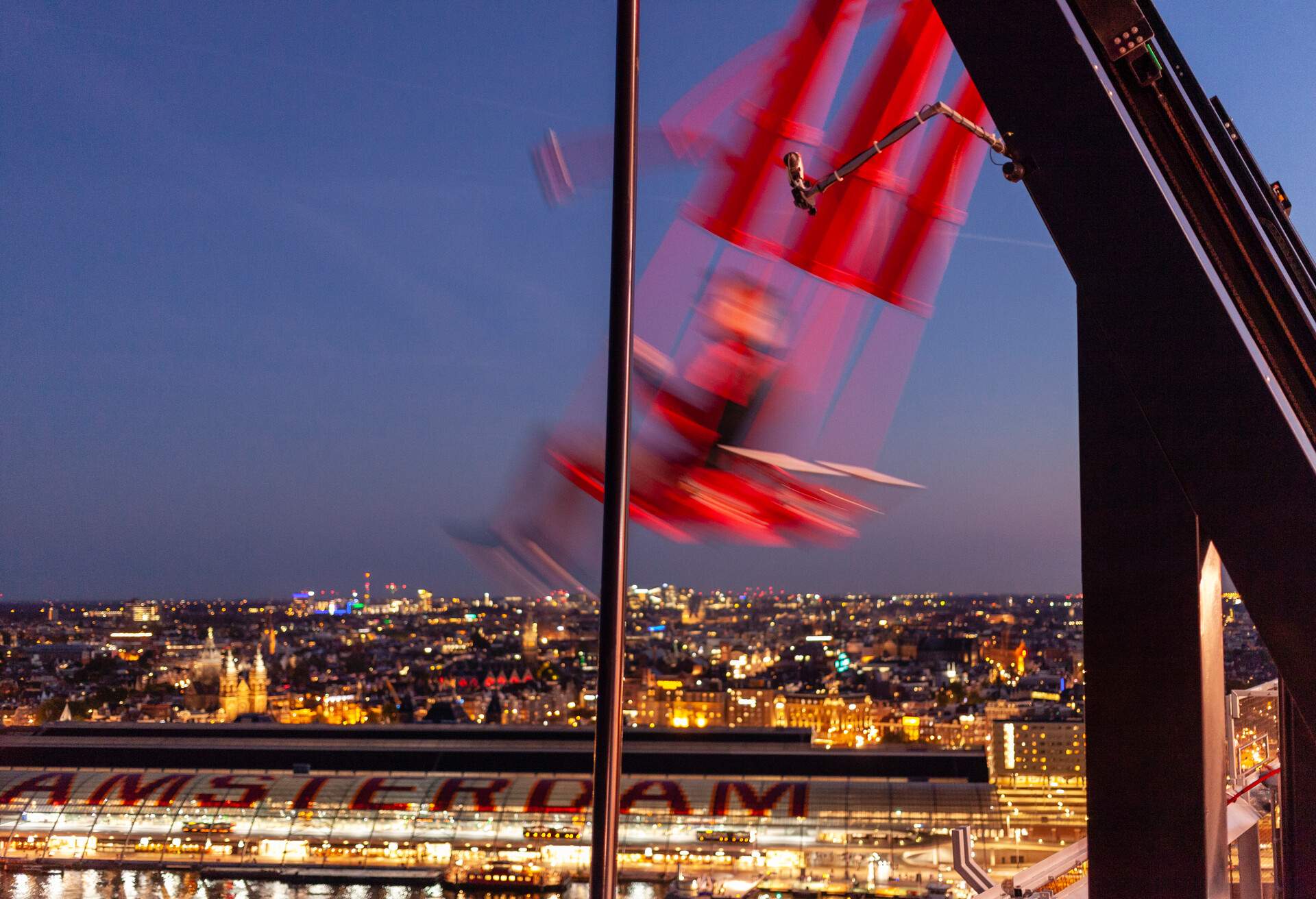 A blurry silhouette of a person on a big red swing with a view of Amsterdam city by night