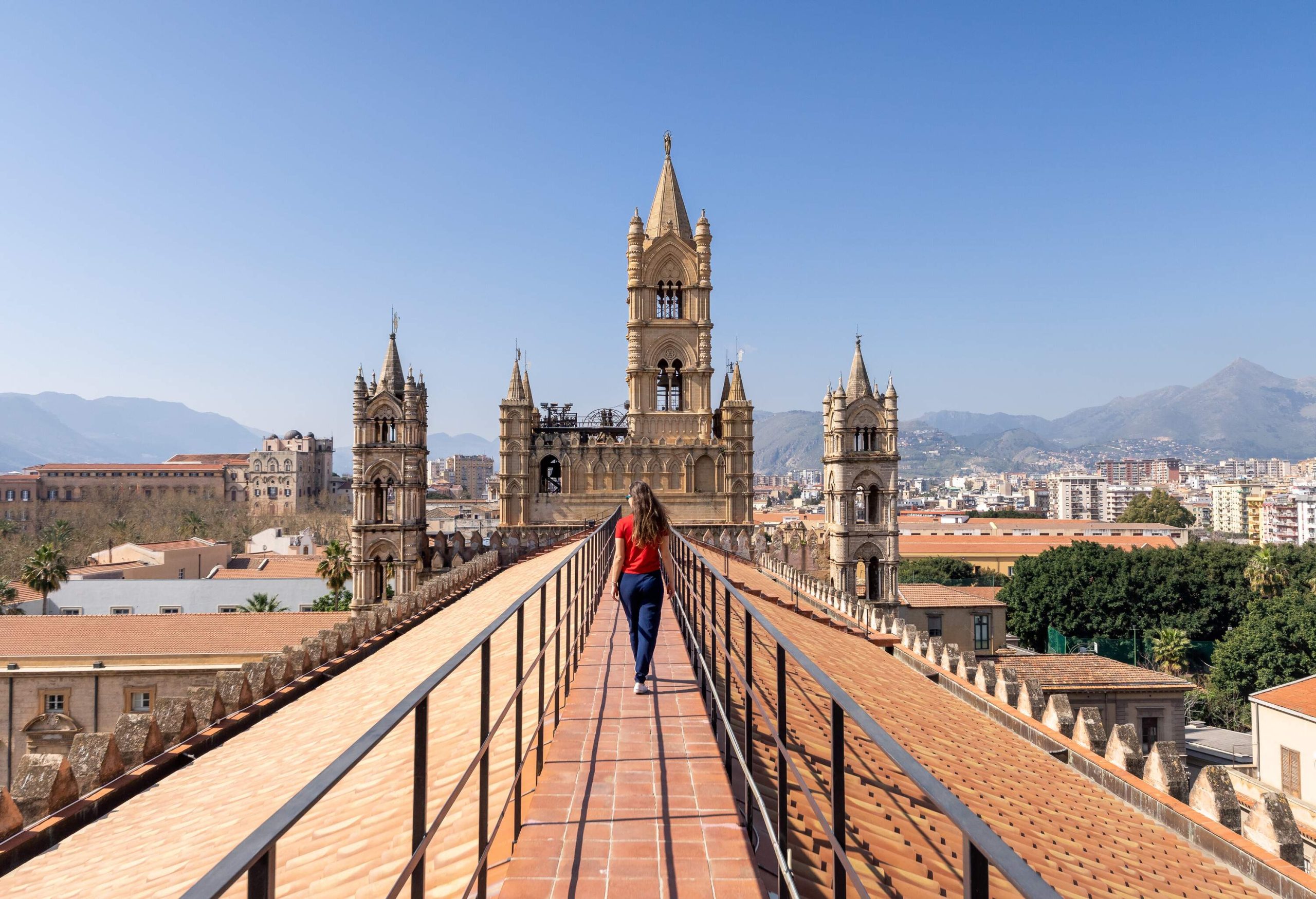 A woman walks on a rooftop walkway with guardrails with a view of the church's towers.