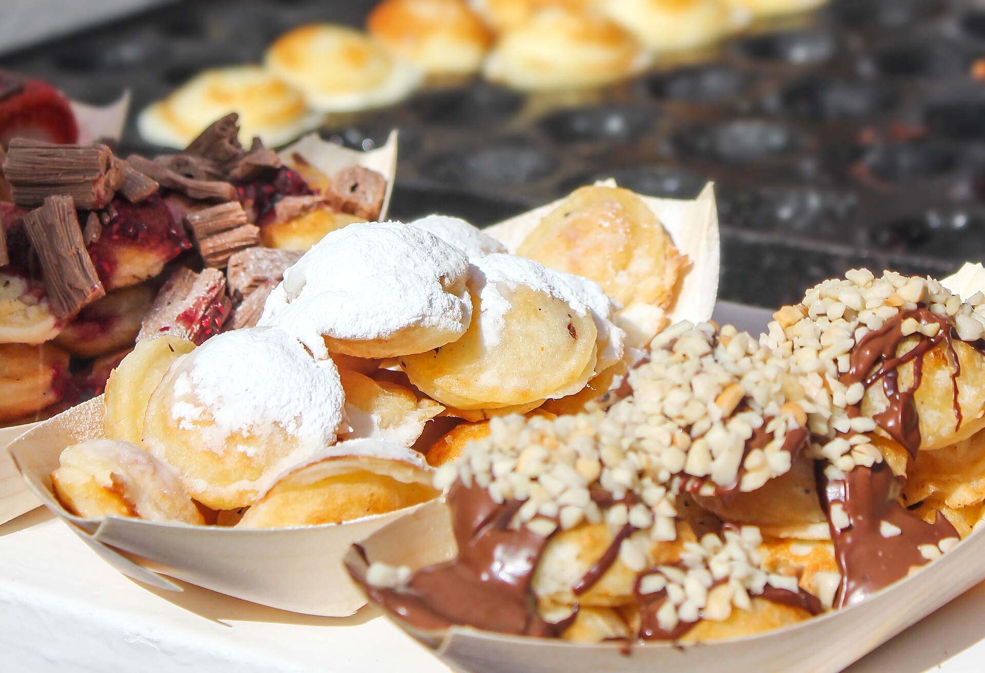 A tempting assortment of sweet Dutch pancakes is adorned with a delightful array of toppings, ranging from a dusting of powdered sugar to a decadent chocolate peanut sauce.