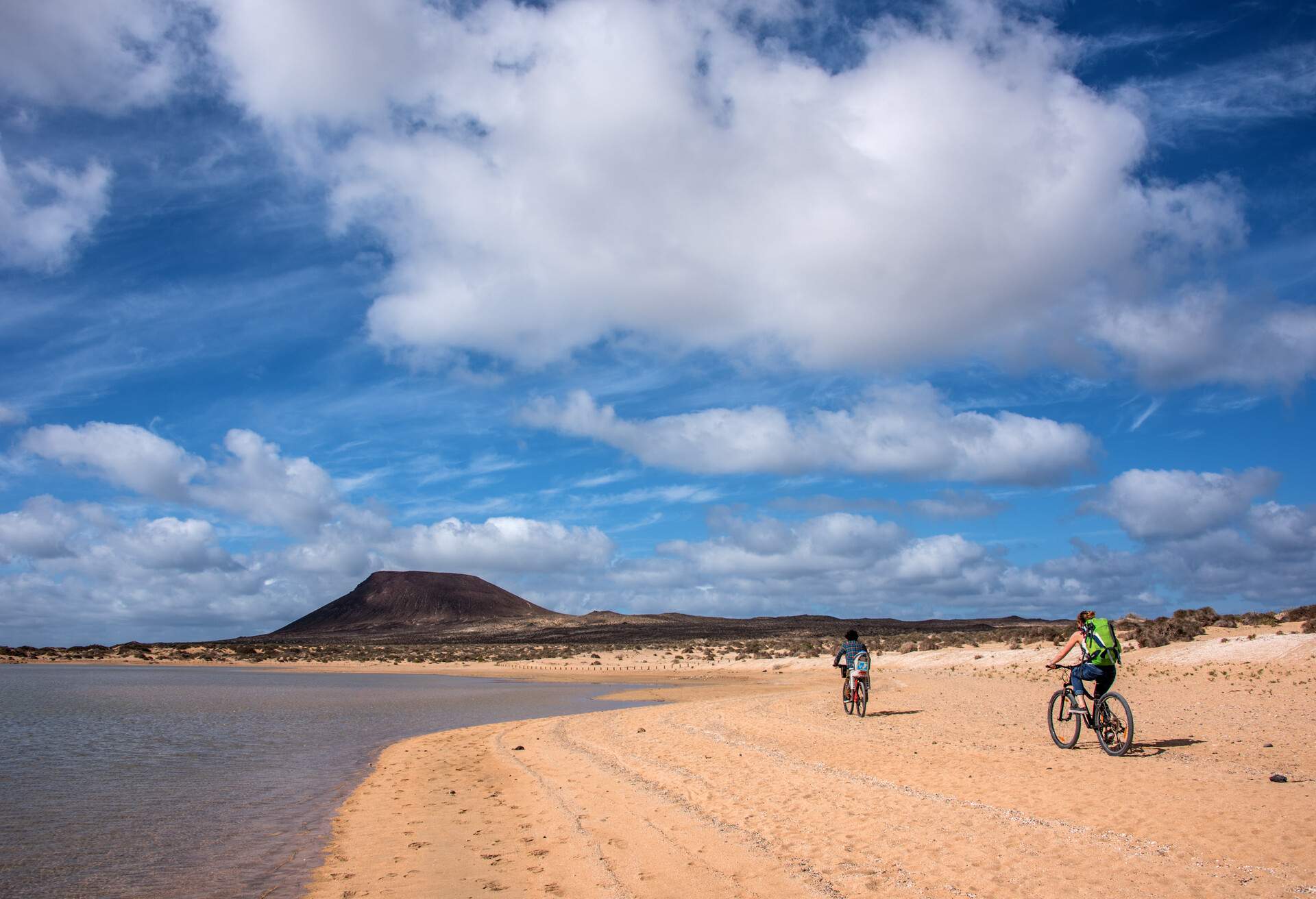 Two people riding their bikes on the sand at the beach with a volcano in the distance.