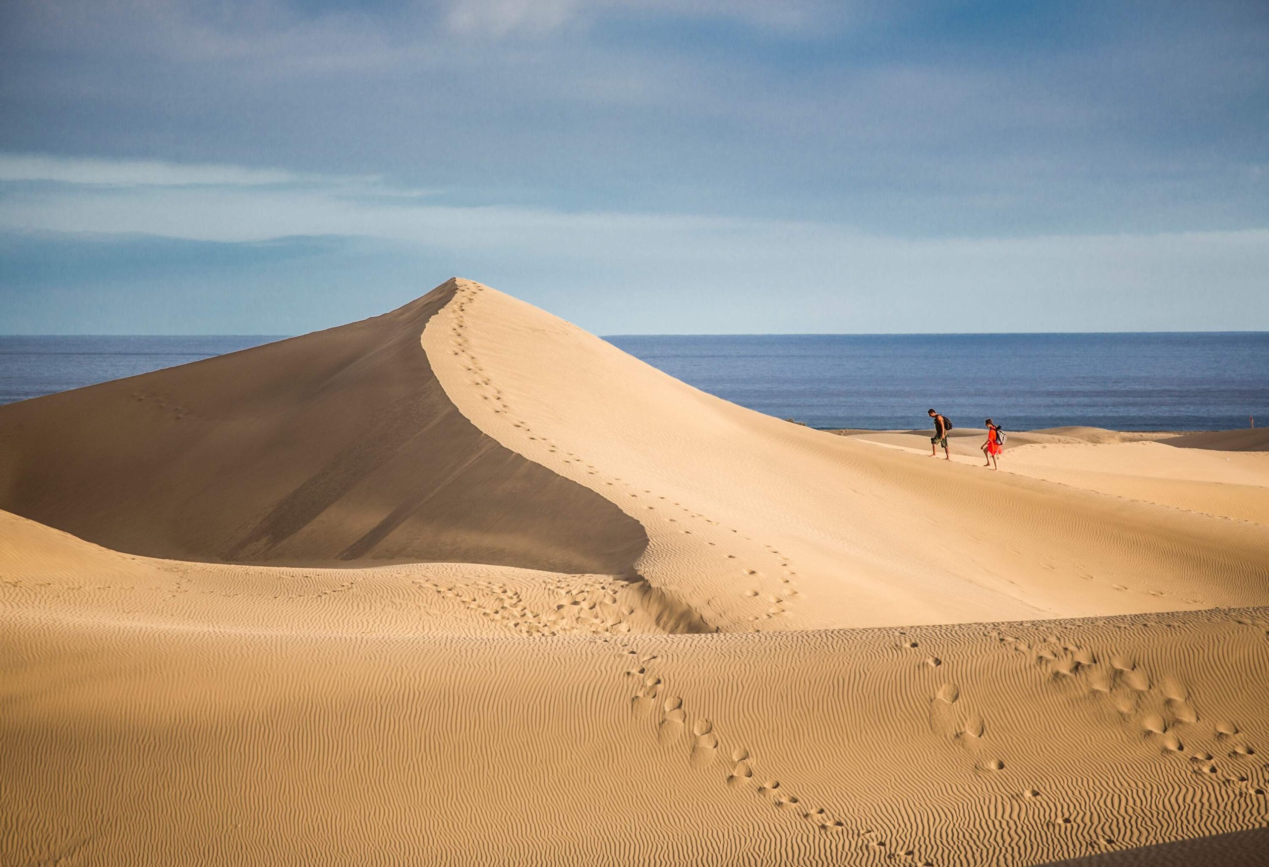 Under an overcast sky, two people are strolling down a sand dune adjacent to the sea.