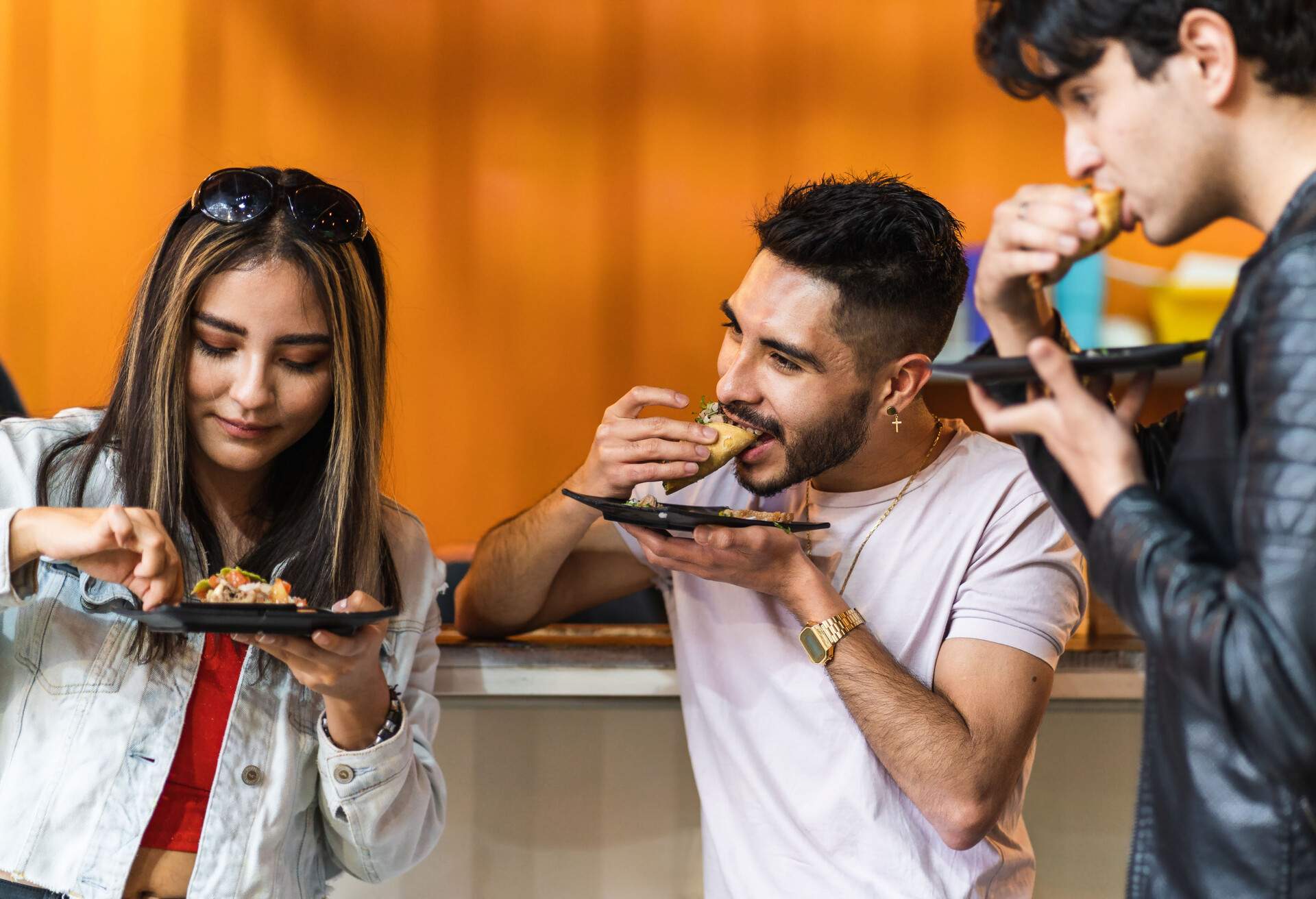 Three friends in casual clothes take a bite of tacos from small plates.