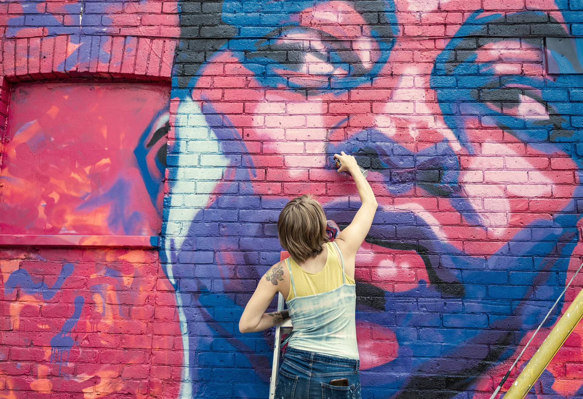 A female artist spray paints a colourful mural on a brick wall.