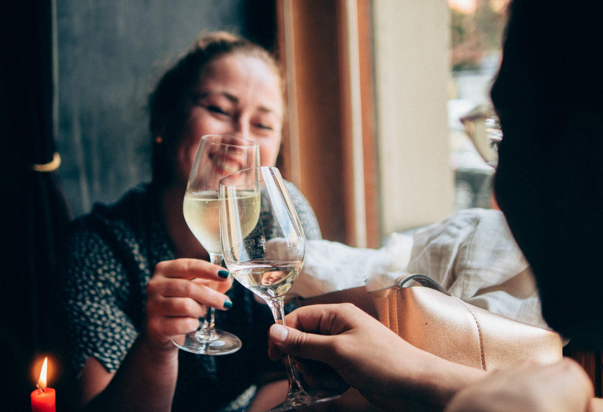 A woman smiling as she clinks her wine glass with the person in front of her.