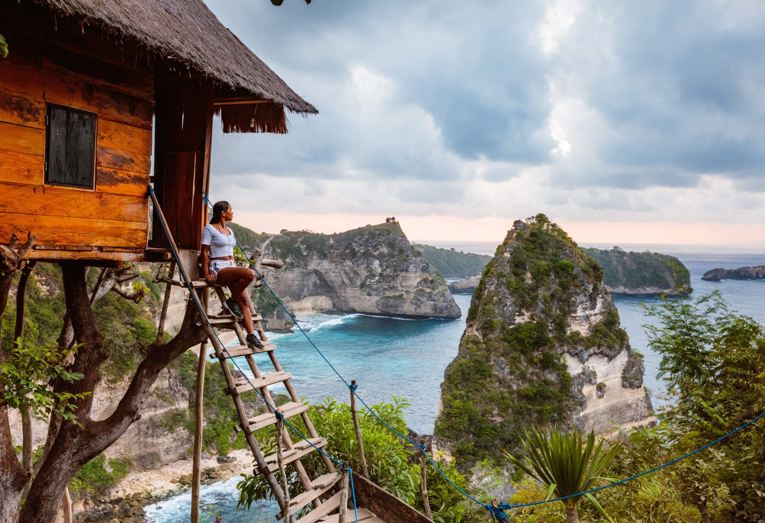 A woman sits on the wooden steps of a wooden tree house and looks out at the lush rocky islands on the blue sea.