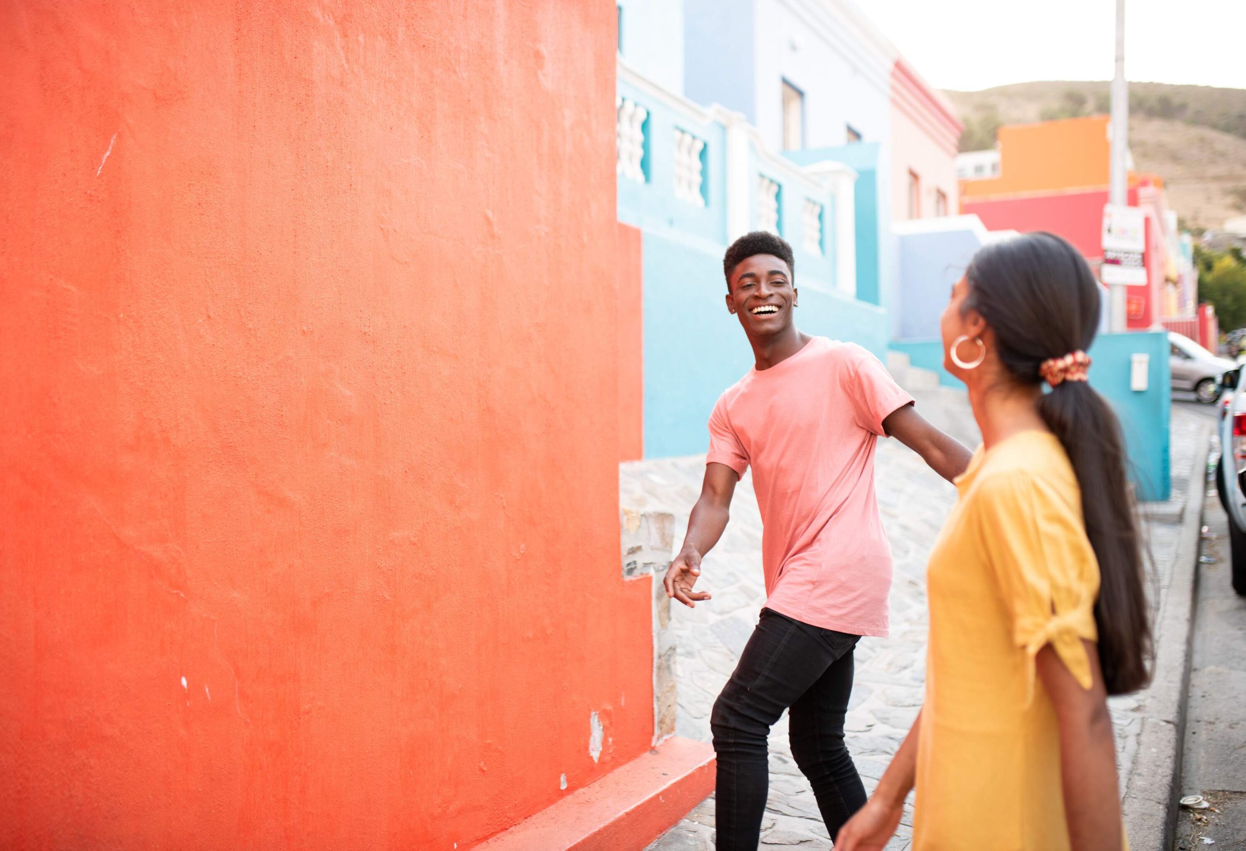 A young cheerful couple standing on the sidewalk bordered with multicoloured building walls.