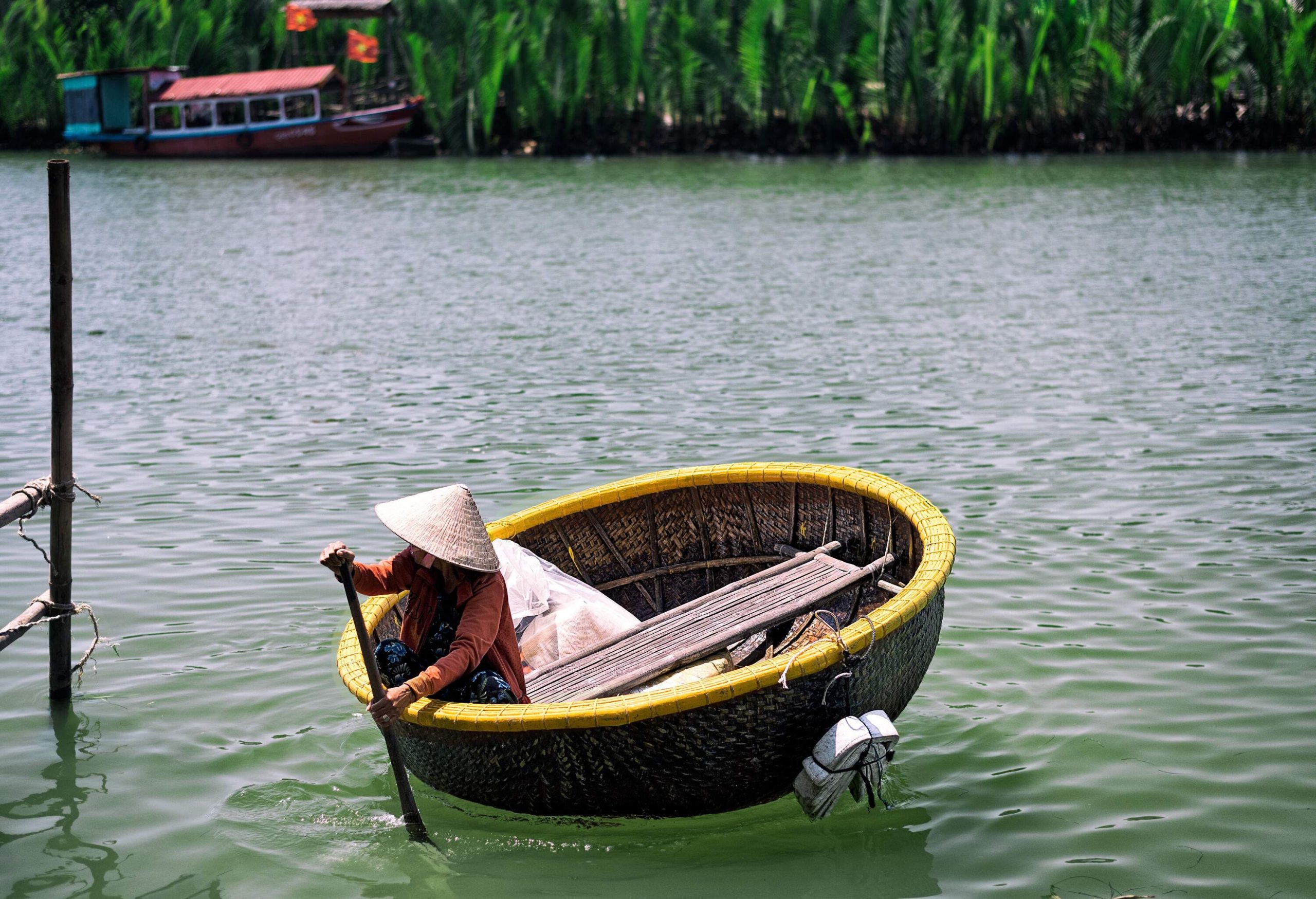 A person in a straw hat rides a circular straw boat and paddles across the river.