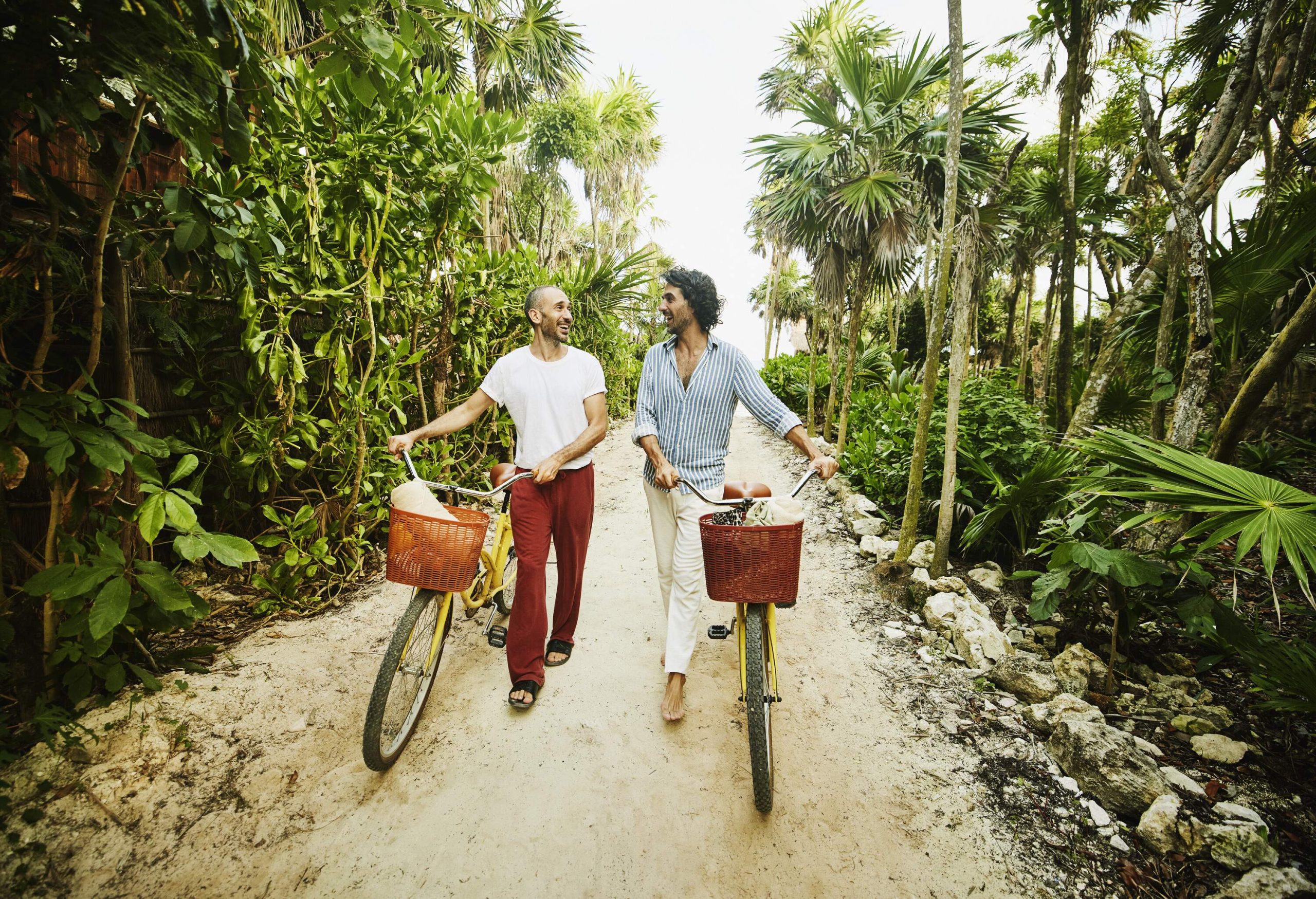 Lgbtq male couple walking on a tropical unpaved road with bicycles amongst trees