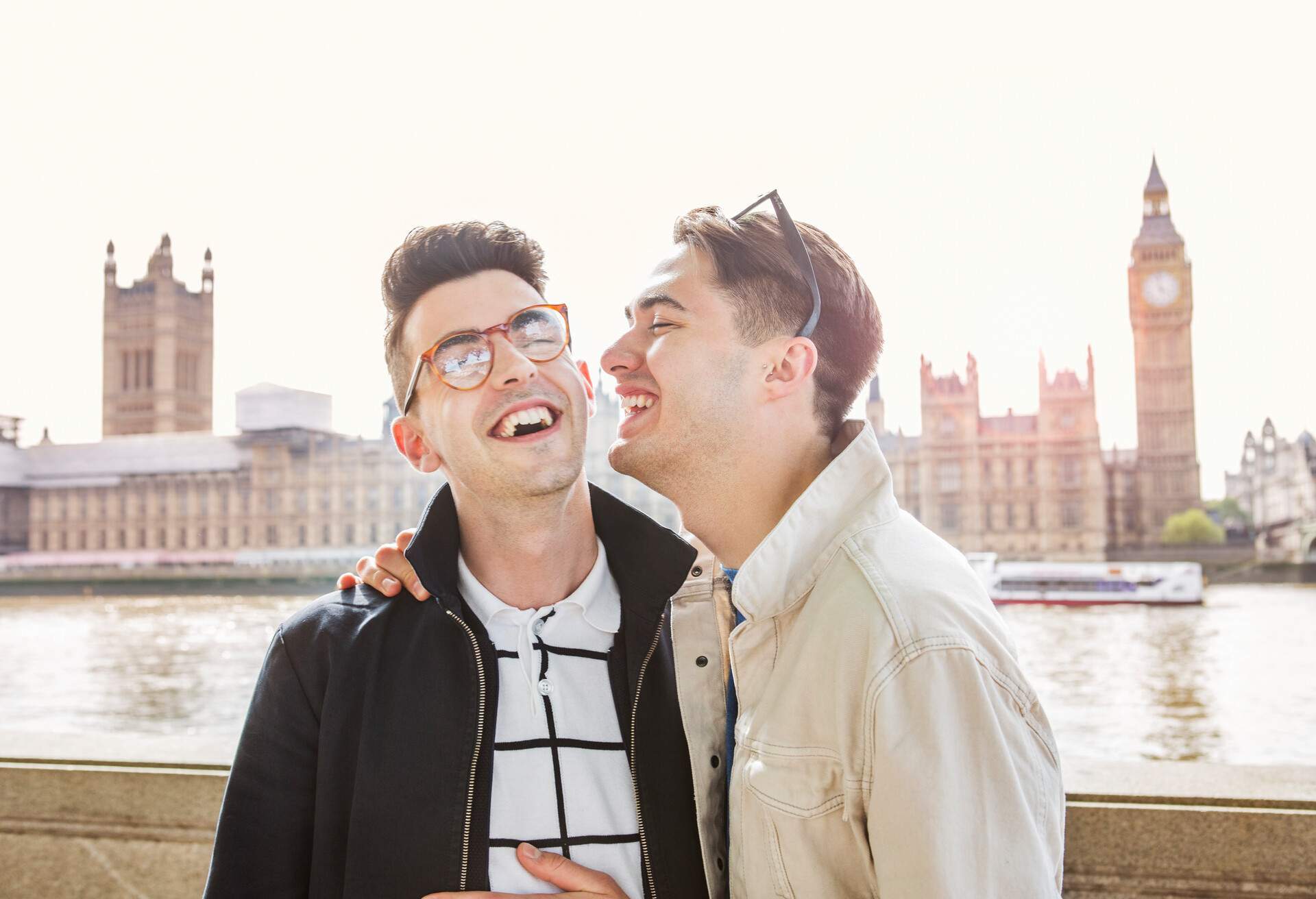 Male couple in black and white jackets laugh while one of them has his arms over the other's shoulder.
