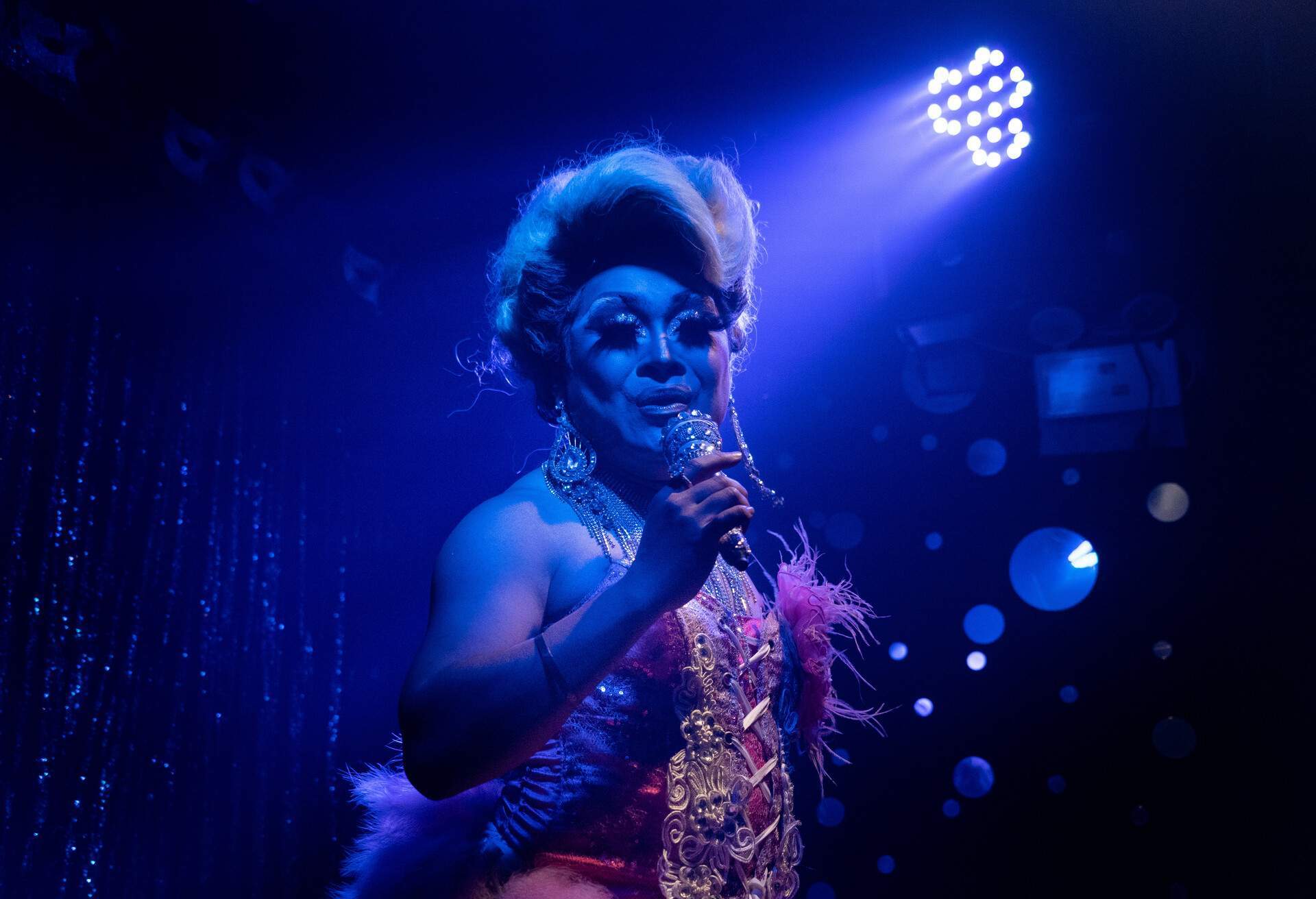 A drag queen illuminated with ultraviolet lights held a microphone while performing.