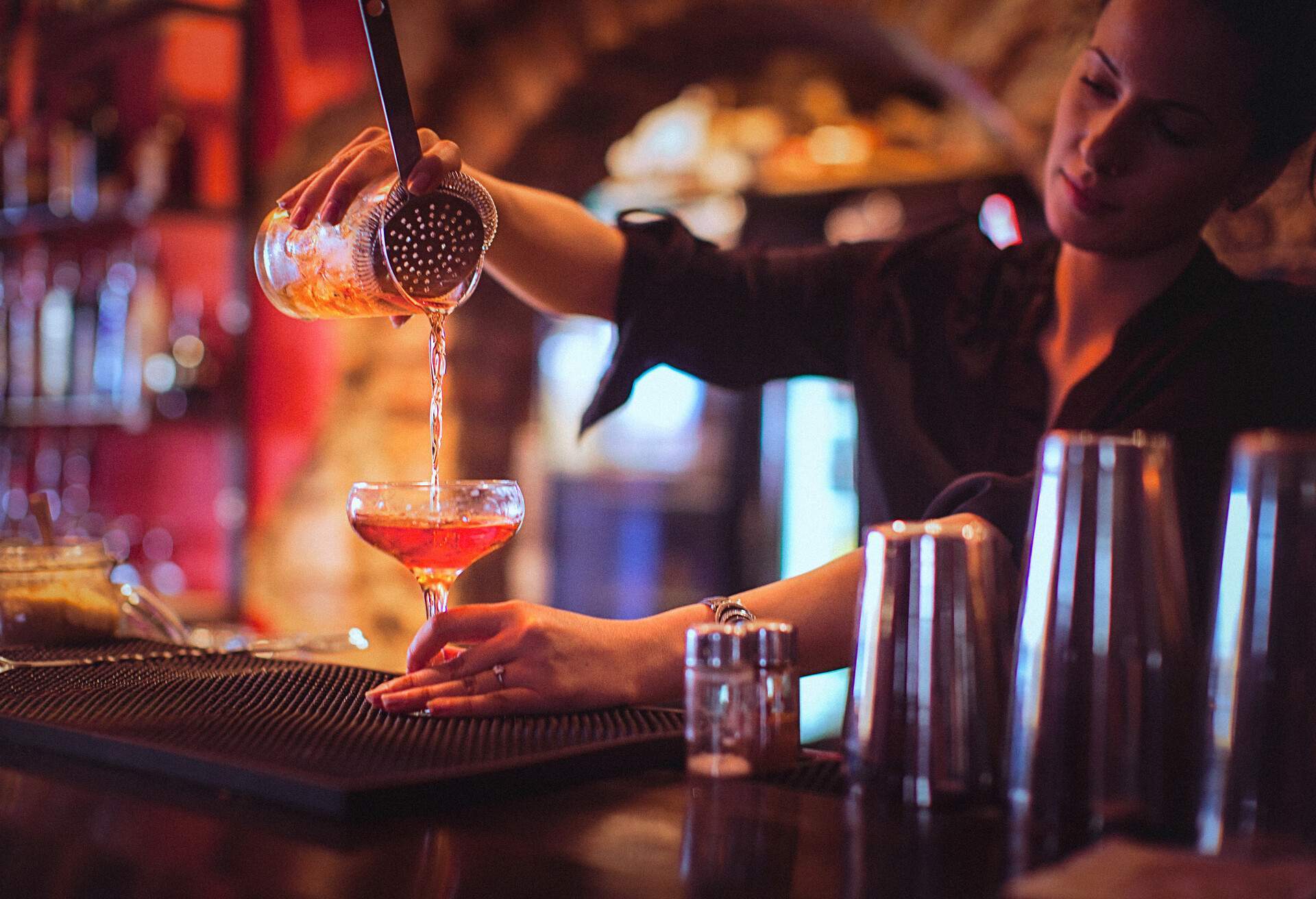 A female bartender pouring a cocktail drink into a coupe glass from behind the counter.