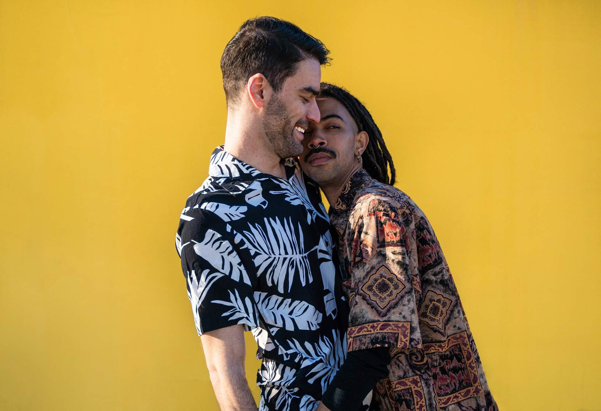 Two male lovers in printed shirts embrace against a yellow backdrop.