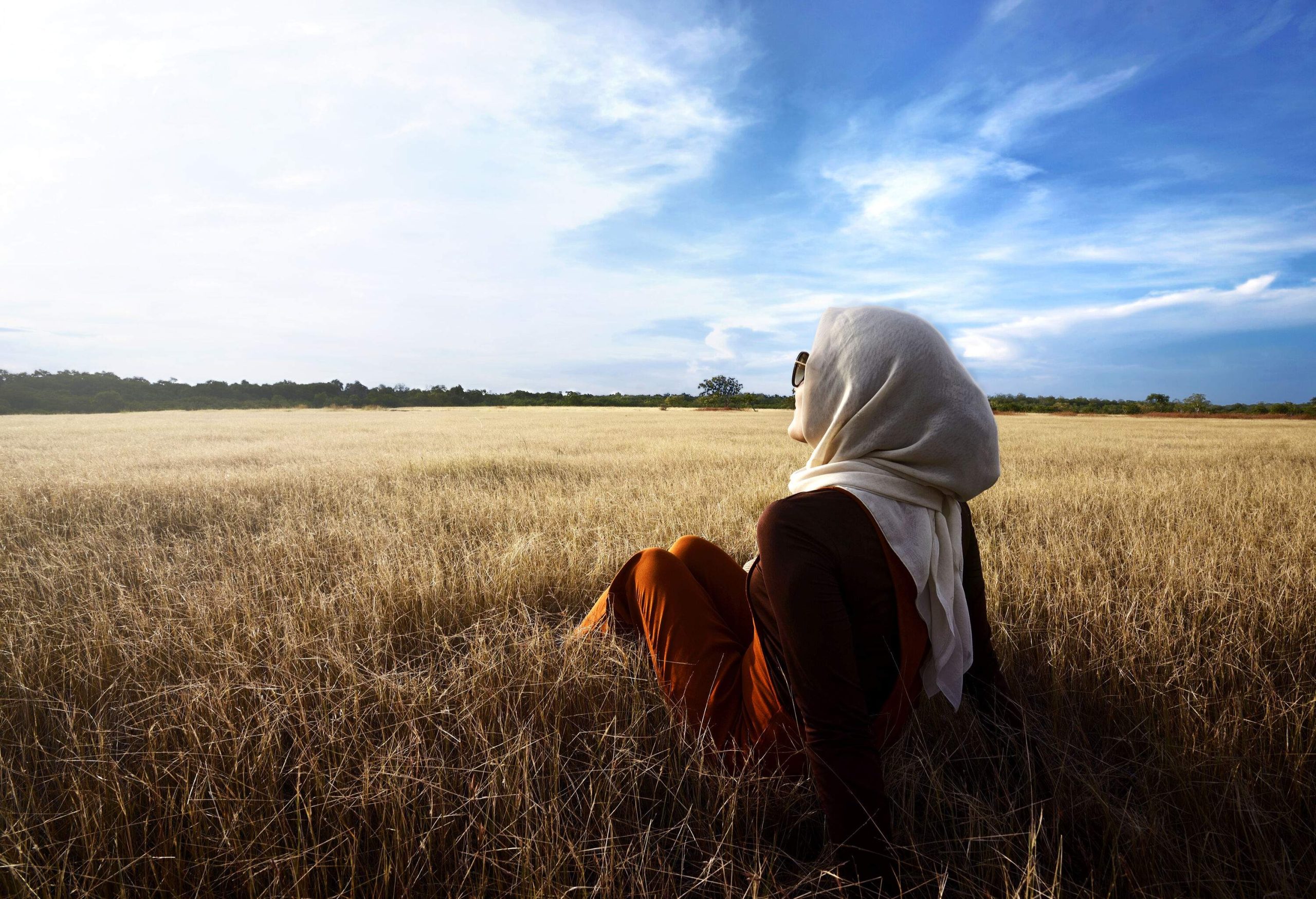 A woman wearing a hijab relaxes on the savanna, soaking up the sun with her sunglasses on as the blue sky stretches out overhead.